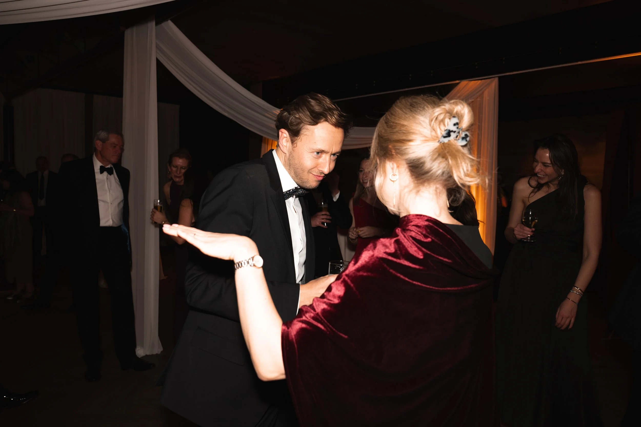 A man and woman dance closely at a formal event, with other guests socializing in the background.