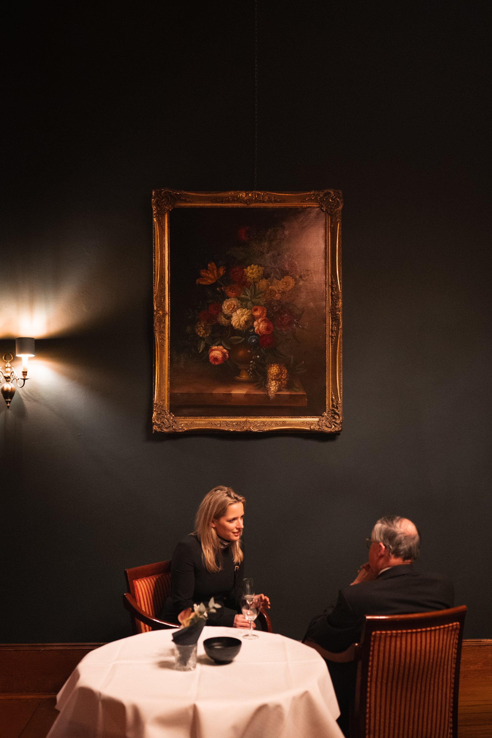 A woman and a man sitting at a table with a white tablecloth in an elegant dimly lit restaurant, talking to each other with a large painting of a flower bouquet hanging on a dark wall behind them.