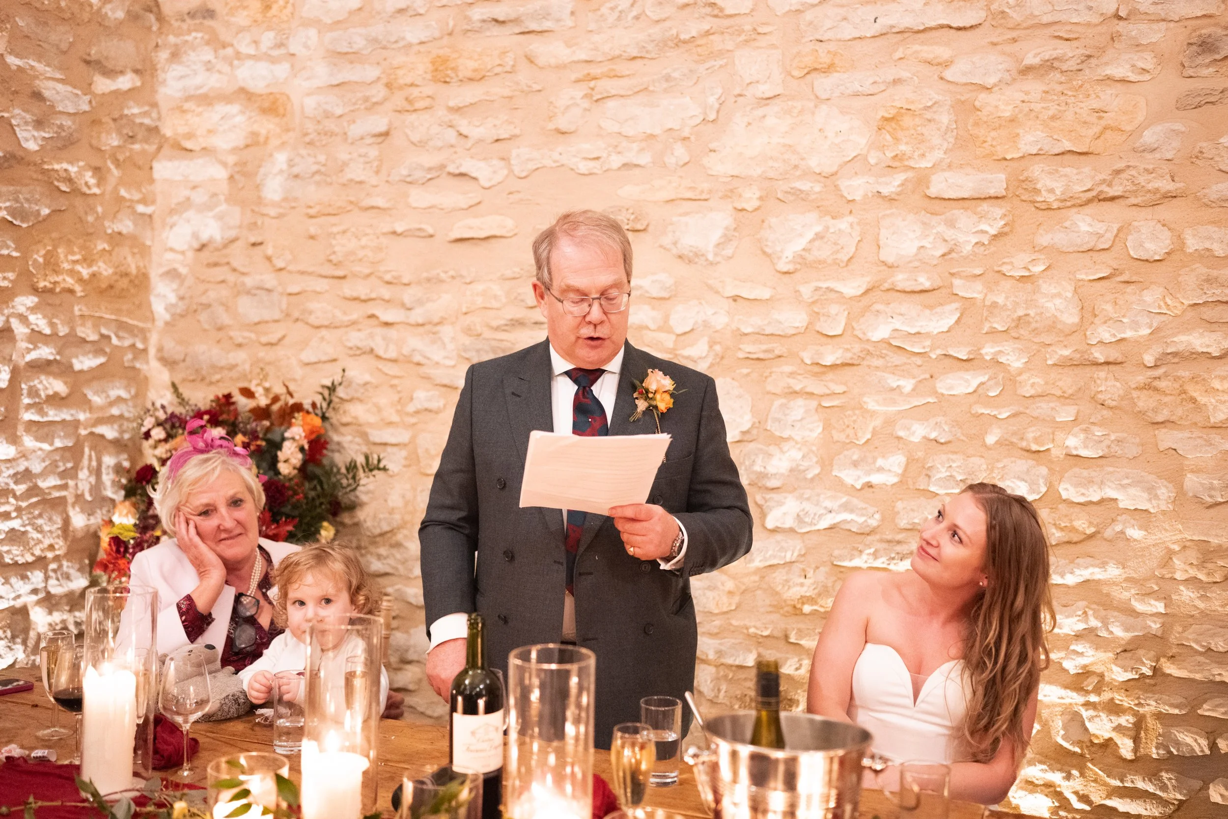 A man gives a speech at a wedding reception, standing at a table with a woman in a white dress and a young girl, while an older woman listens nearby. The table has candles, wine bottles, and floral decorations, and the background features a stone wal
