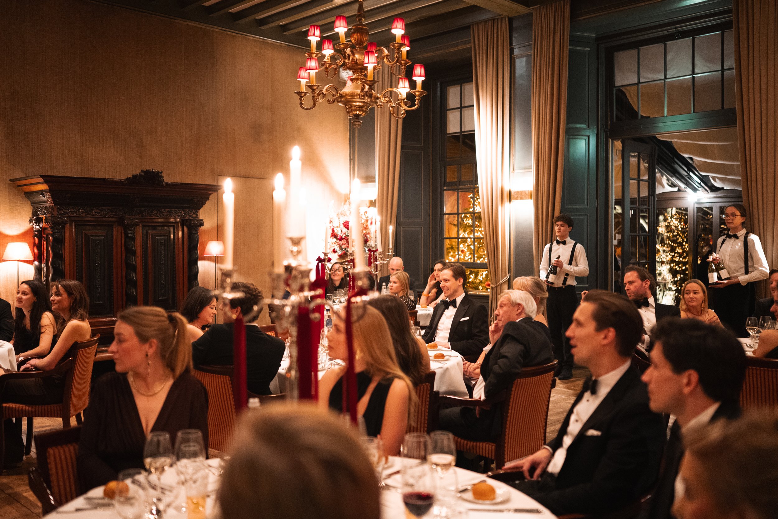 People seated at tables in a formal dining room, dressed in black-tie attire, during a festive event with Christmas decorations and a chandelier hanging from the ceiling.