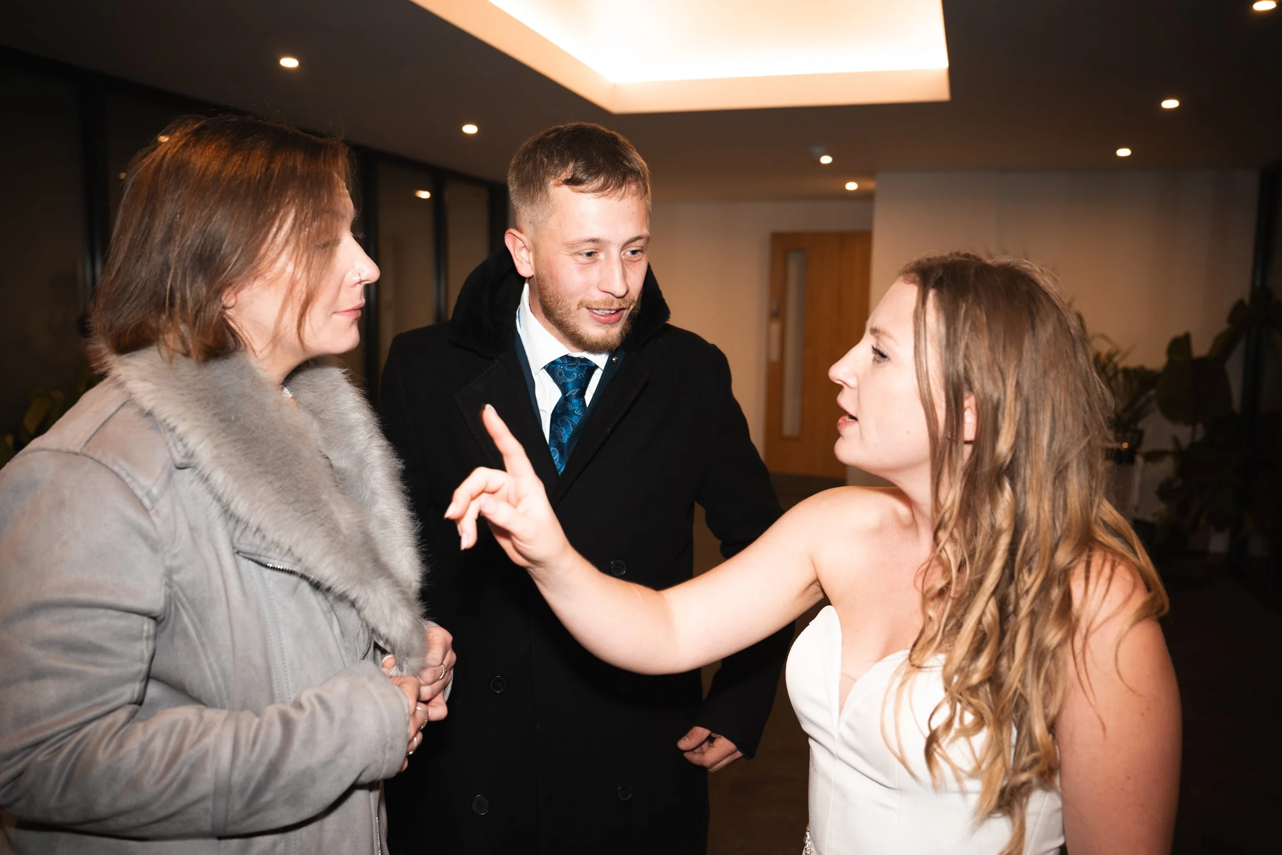A woman in a white dress having a conversation with two other women at an indoor event, with one woman wearing a beige coat with a fur collar and the other dressed in a black coat and tie.