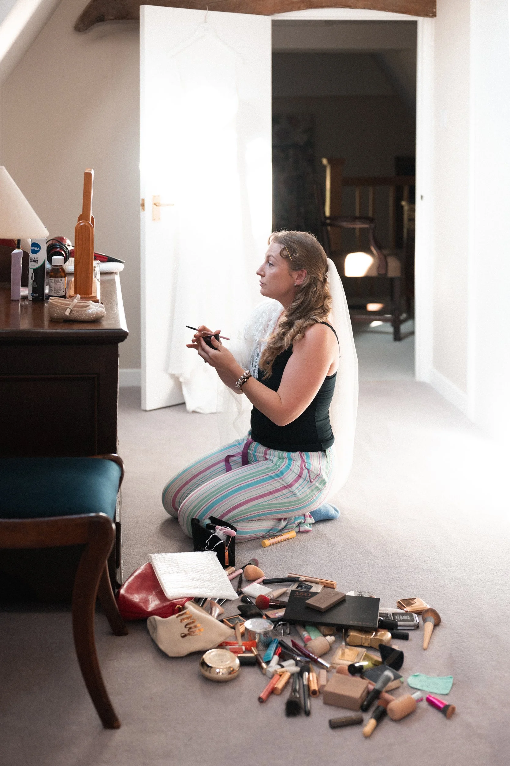 A woman in striped pajama pants and a black tank top kneels on the carpeted floor, applying makeup with an array of makeup products scattered in front of her. She is in a room with a dresser and a partially opened door leading to another space.