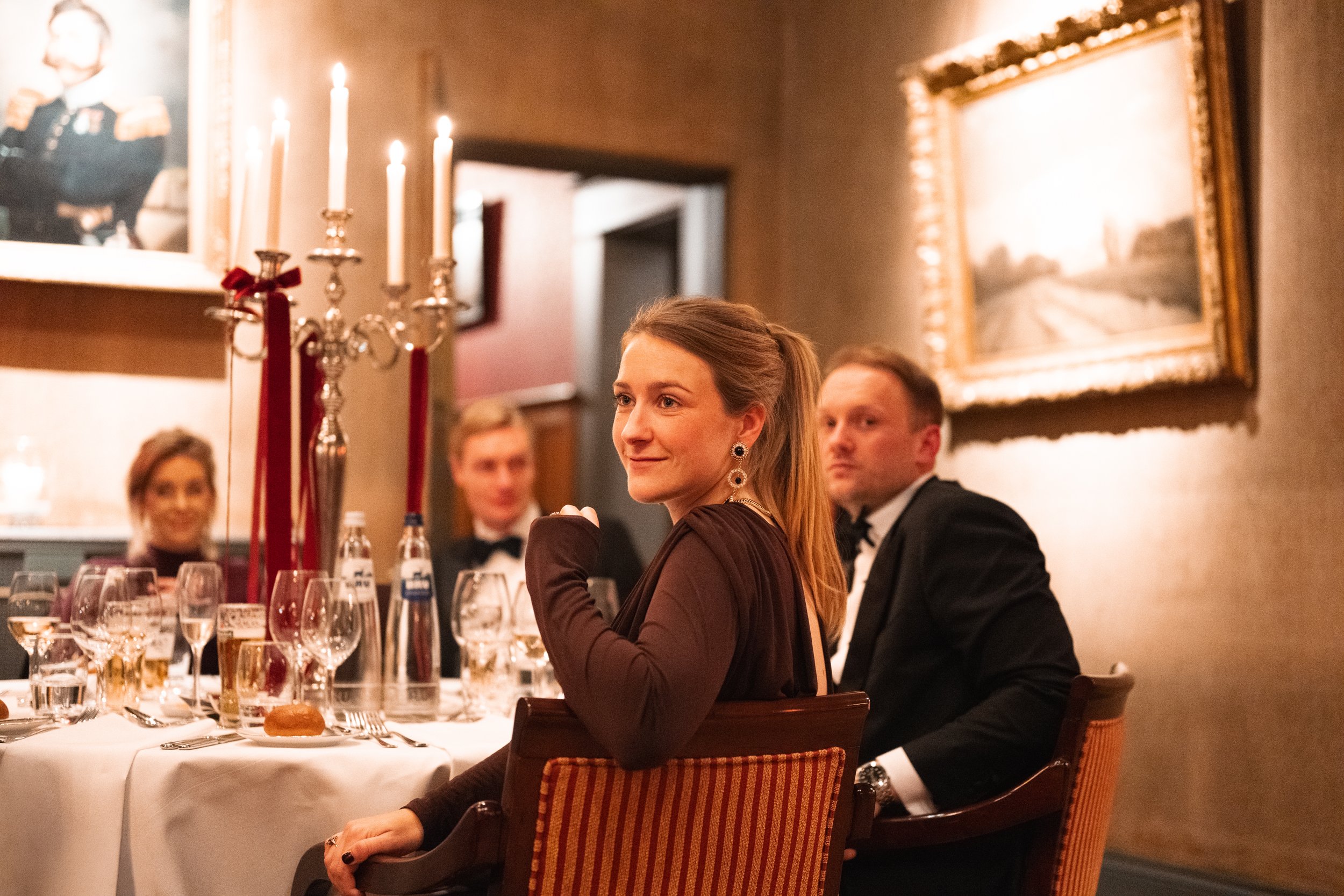 A woman in a brown dress with earrings sitting at a formal dinner table with three men dressed in tuxedos; the table has glasses, bottles, and a candelabrum with lit candles; the setting appears to be an elegant restaurant or banquet hall.
