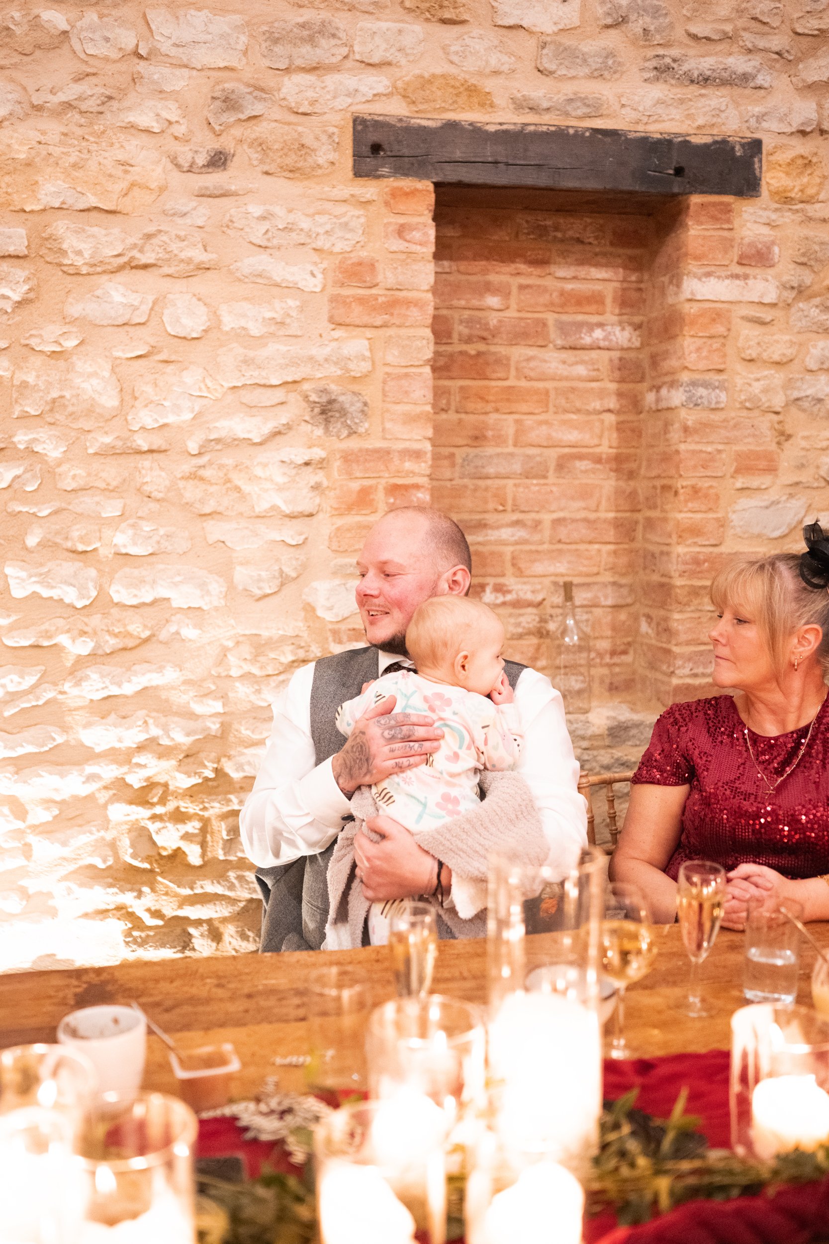 A man holding a baby, sitting next to a woman, at a decorated table with candles, in front of a textured brick wall.