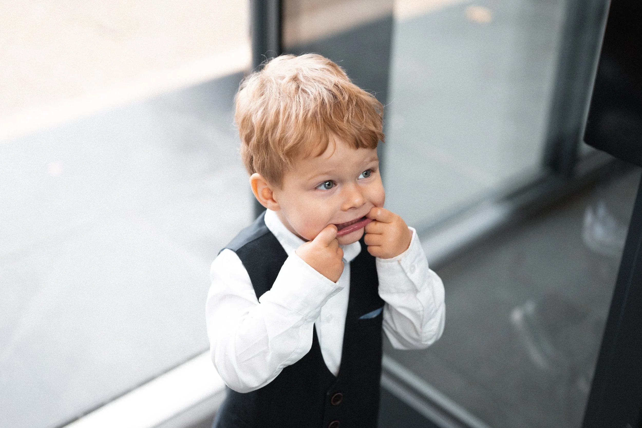 A young boy with red hair and blue eyes making a funny face by pulling at his cheeks with his fingers, wearing a white shirt and a dark vest, standing inside a building near glass doors.