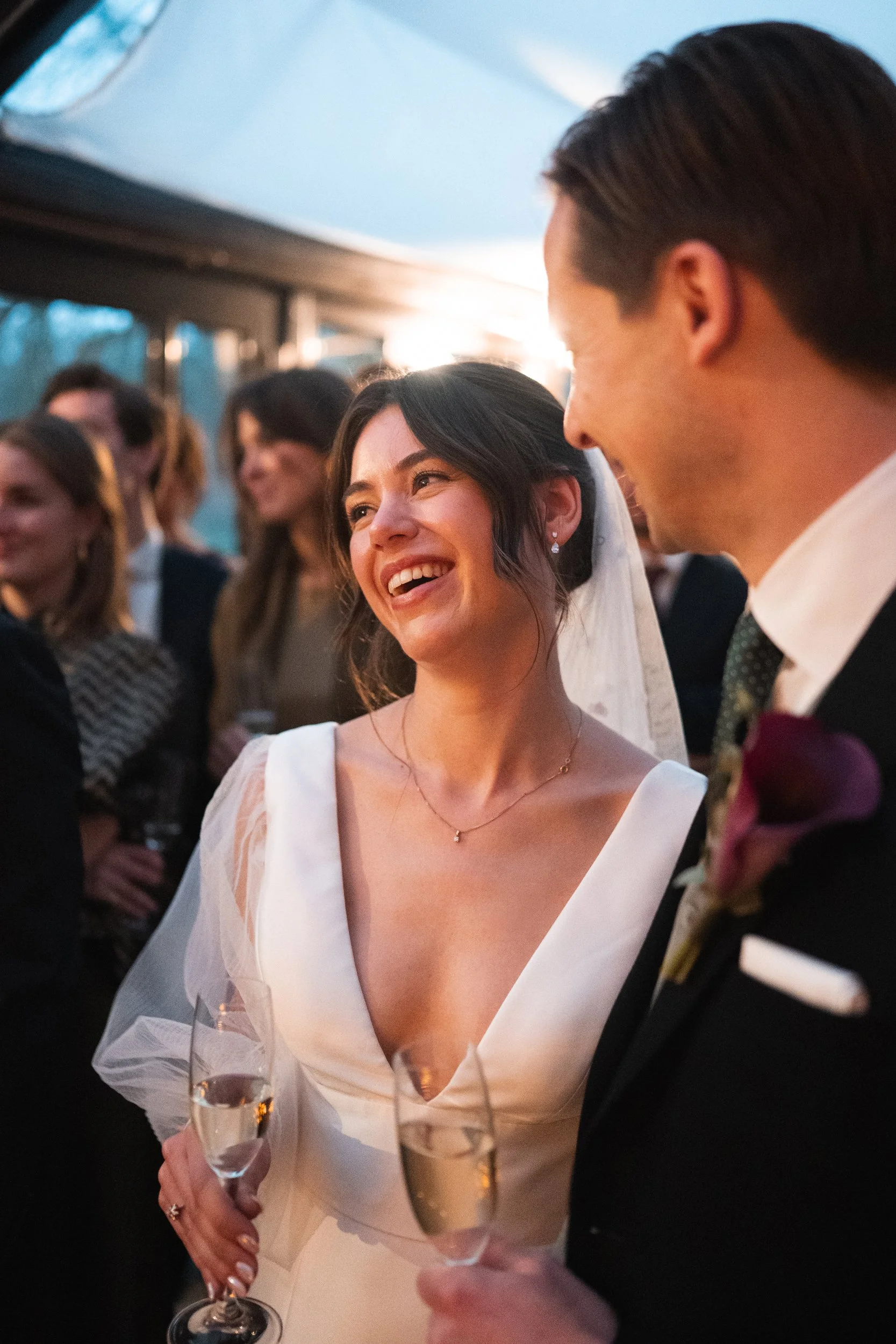 Bride and groom at their wedding reception, smiling and holding champagne glasses, surrounded by guests.