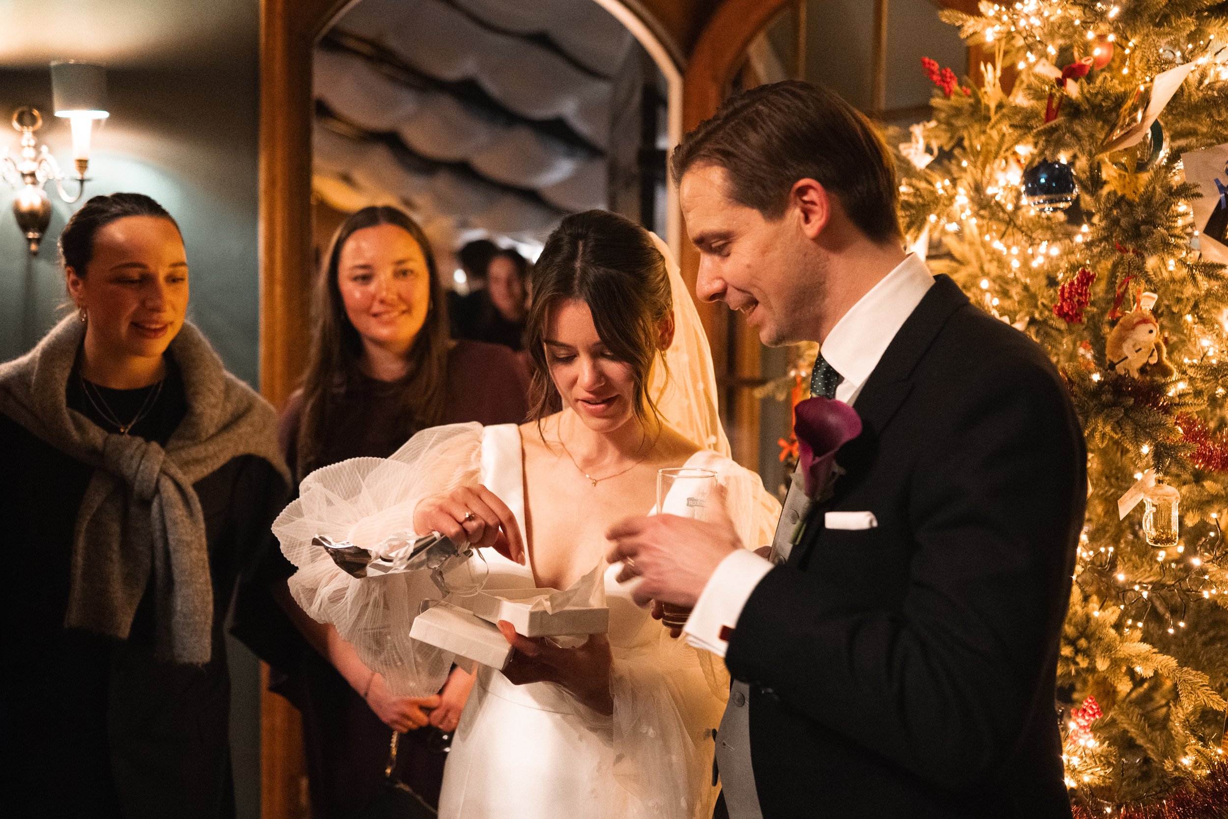 A bride and groom exchange gifts at a Christmas celebration, surrounded by friends and a decorated Christmas tree with lights and ornaments.