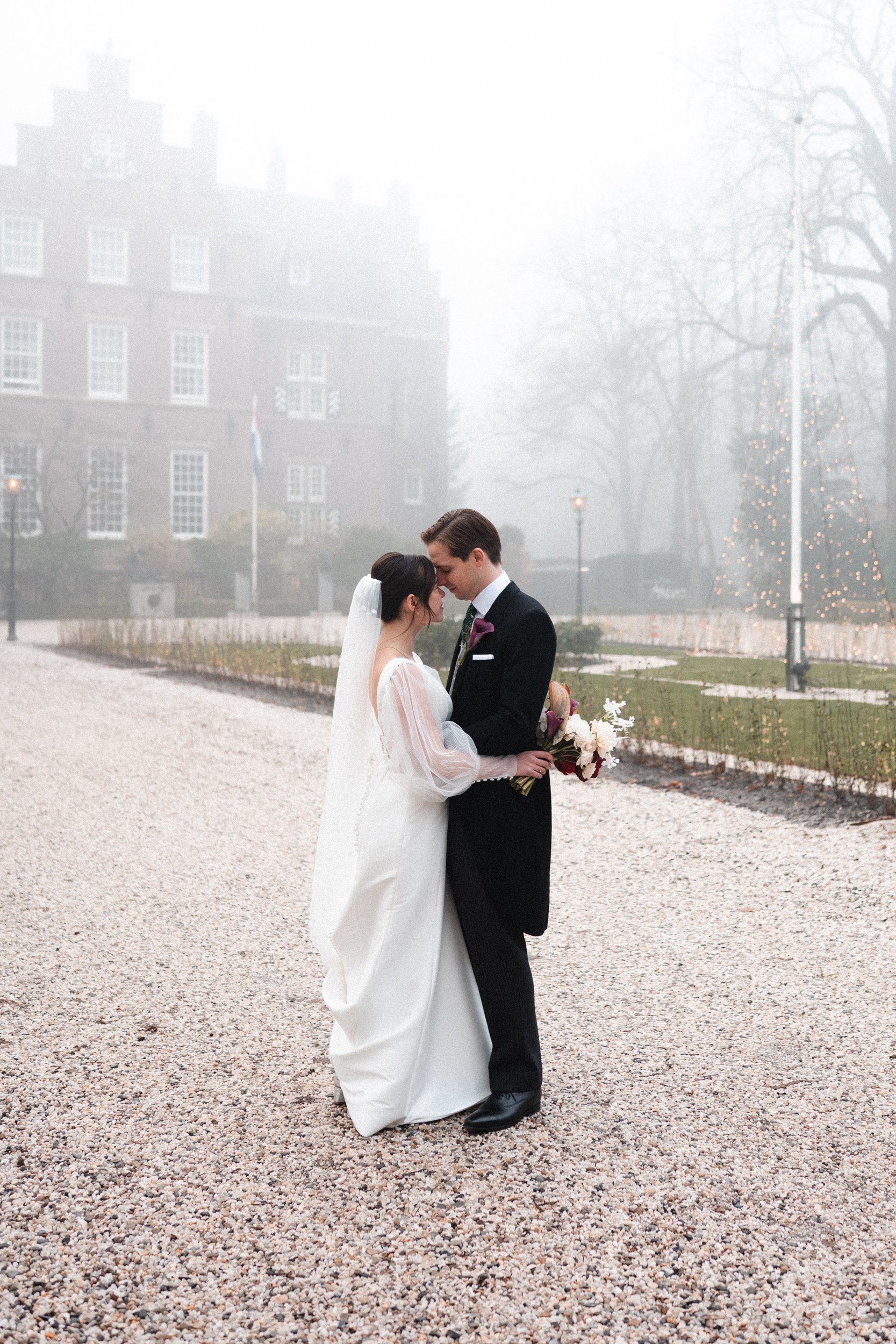 A bride and groom sharing a tender moment outdoors on their wedding day, with foggy weather and a Christmas tree decorated with lights in the background.