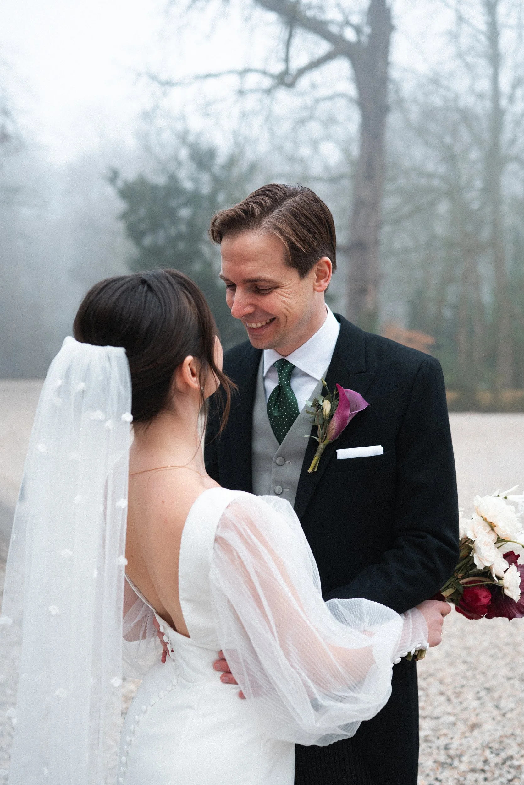 A bride and groom share a moment outdoors on a foggy day, with the groom smiling and holding a bouquet of flowers.