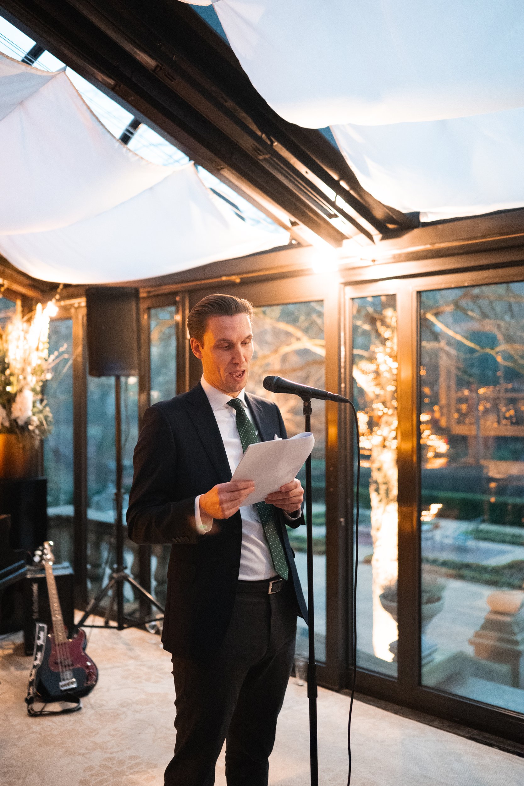 A man in a suit with a green tie speaking into a microphone while reading from papers at a formal event in a room with large windows and white drapery.