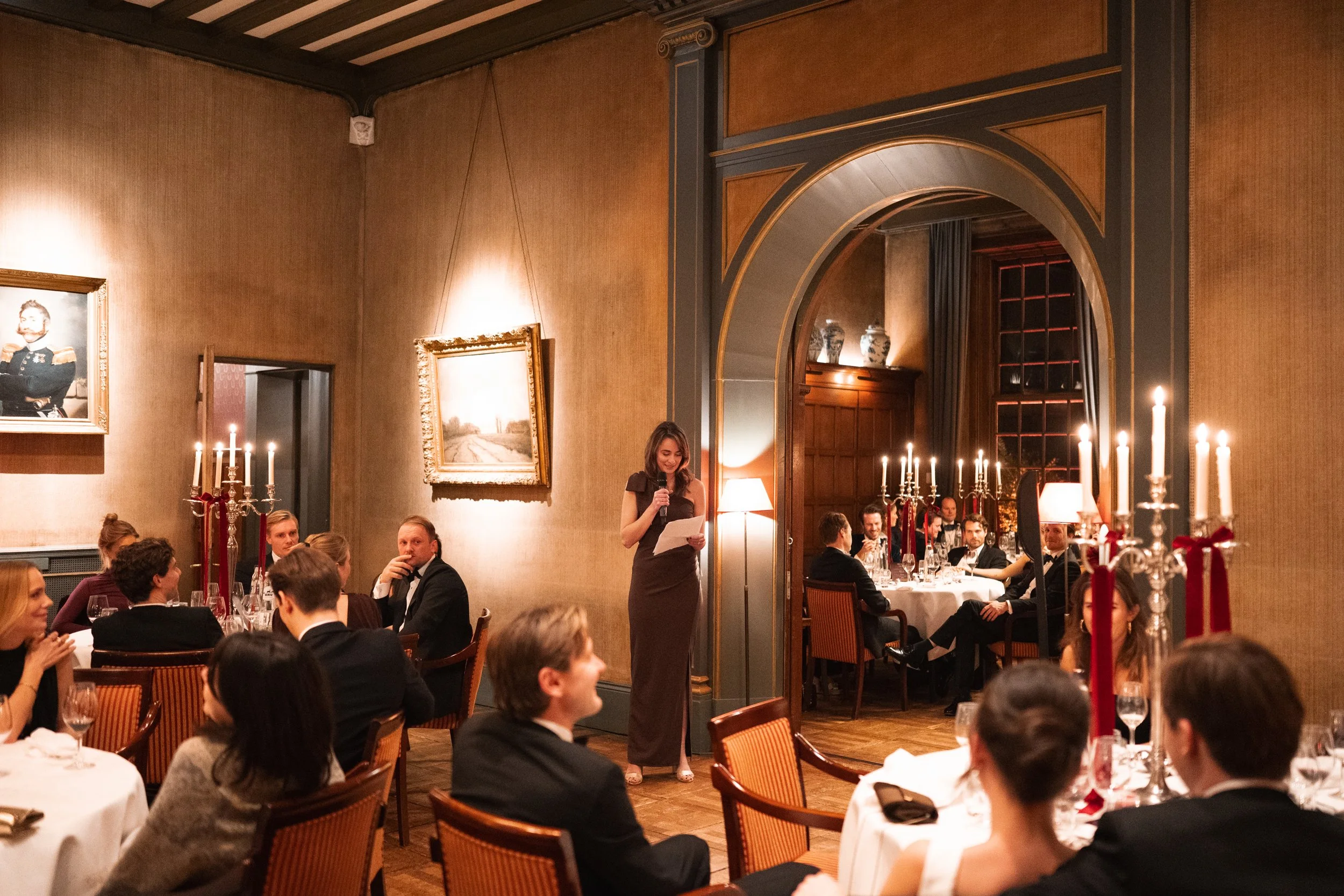 A woman in a dark dress giving a speech at a formal dinner event in an elegant room with candelabras and framed artwork on the walls. Guests seated at round tables watch her.