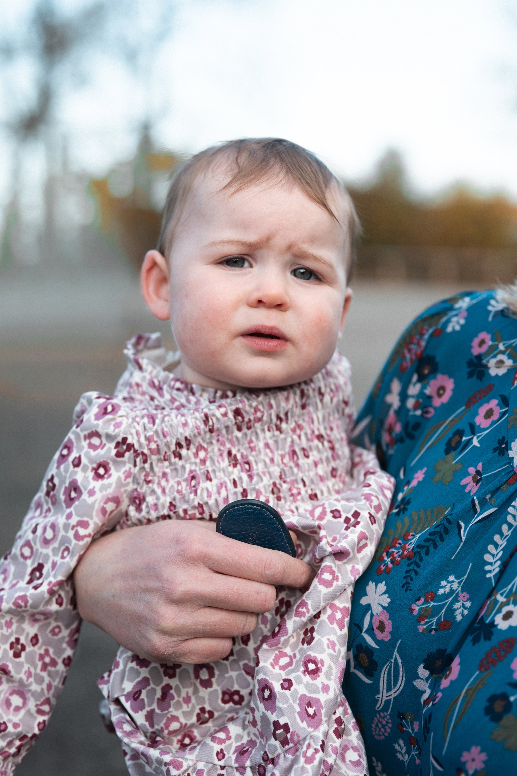 A young girl in a floral dress being held by an adult, with a concerned expression on her face, outdoors in a park or playground area.