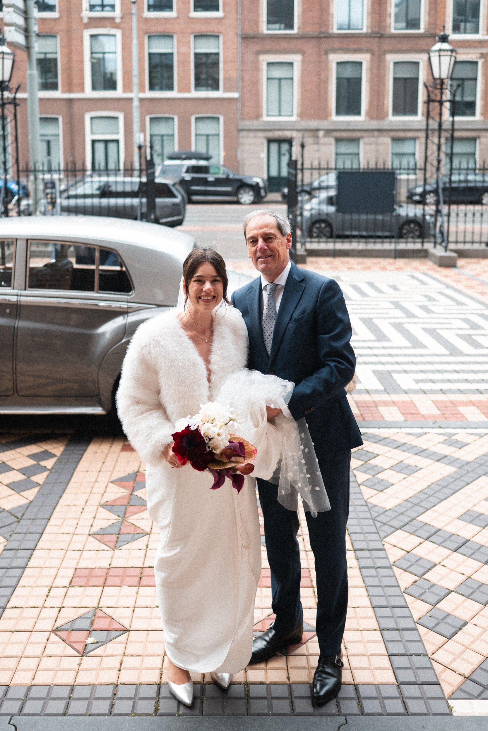 A bride and a man, possibly her father, standing outside on a patterned tiled sidewalk in front of a vintage car, smiling. The bride holds a bouquet of flowers and wears a white dress with a fluffy white jacket. The man wears a navy blue suit with a 