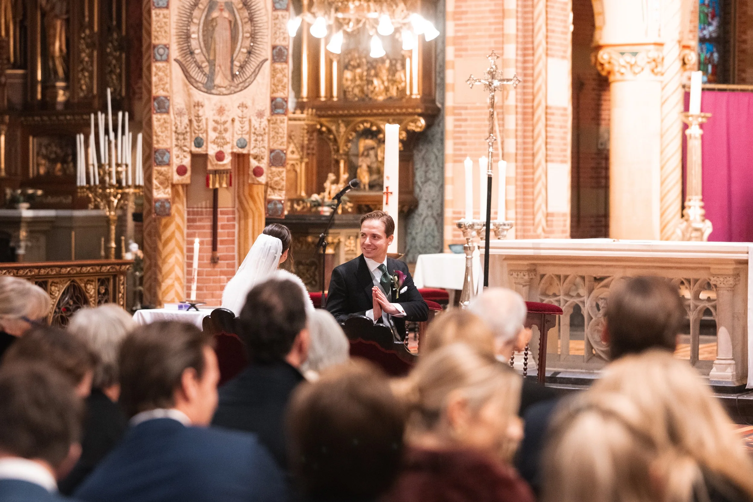 A wedding ceremony inside a church, with a bride and groom seated at the altar. The groom is smiling with hands together in a prayer-like gesture, while the bride, wearing a veil, faces him. The congregation is seated in front, watching the ceremony.