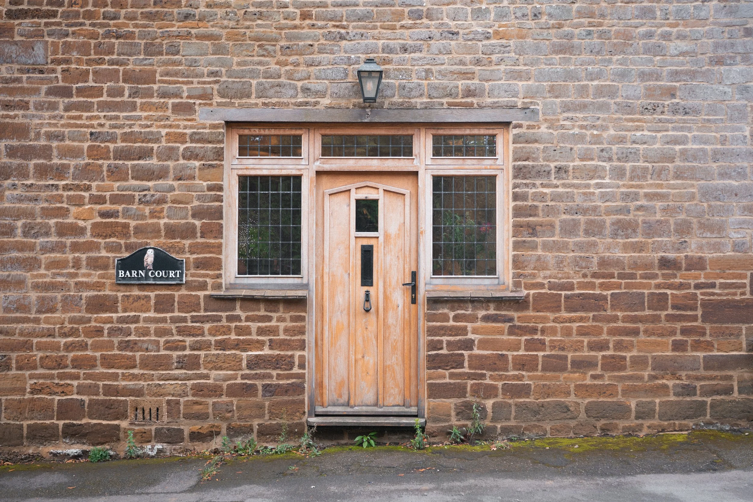 Wooden door with a small window and black handle, set into a brick wall, with two large glass-paneled windows on either side, and a black lantern above. A black sign reads 'Barn Court' with a picture of an owl.