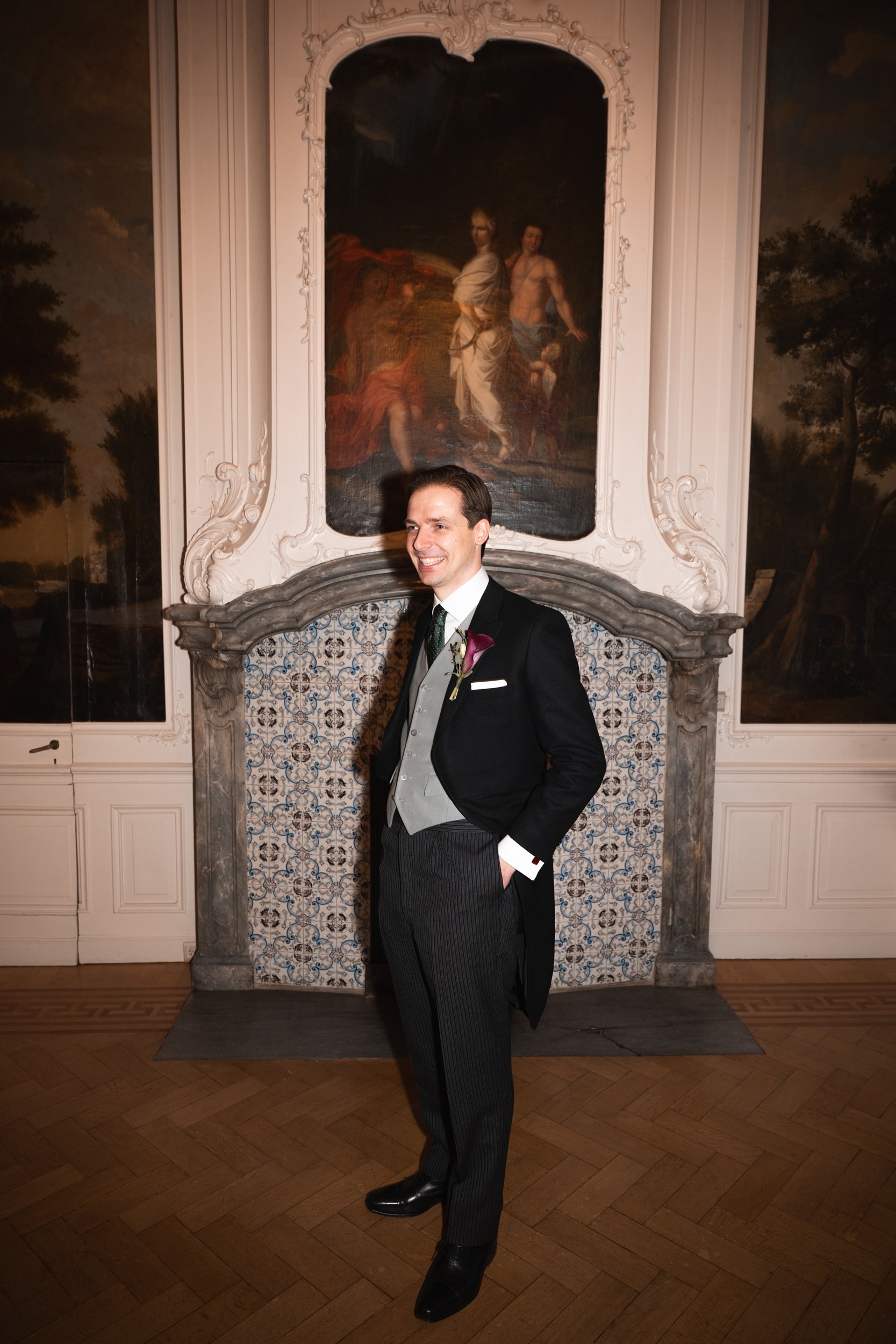 A man dressed in a formal tuxedo with a boutonniere stands smiling in front of an ornate fireplace with a ancient painting above it, inside a decorated room.