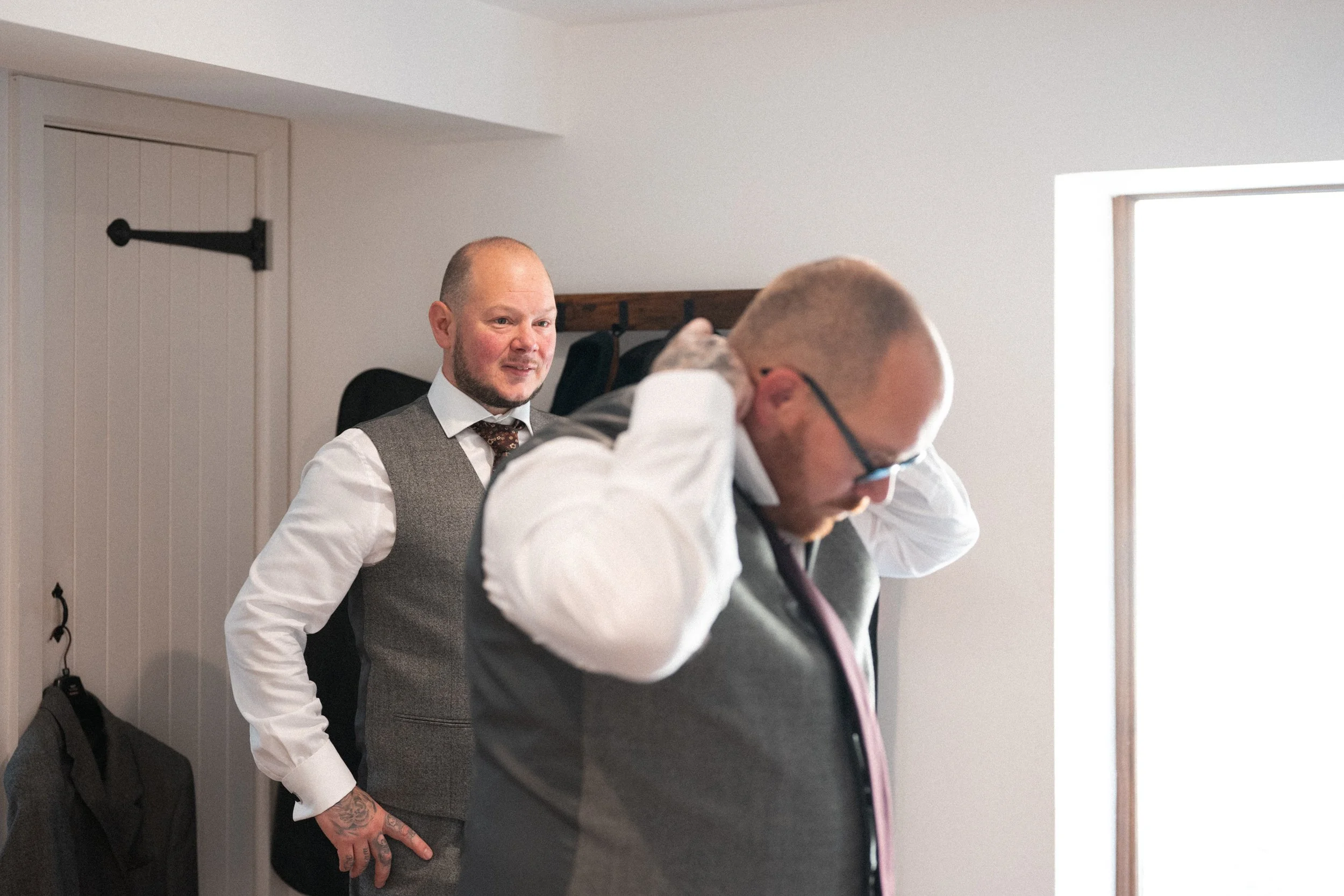 Two men in formal attire, one adjusting his tie and the other standing nearby, in a room with natural light coming through a window.