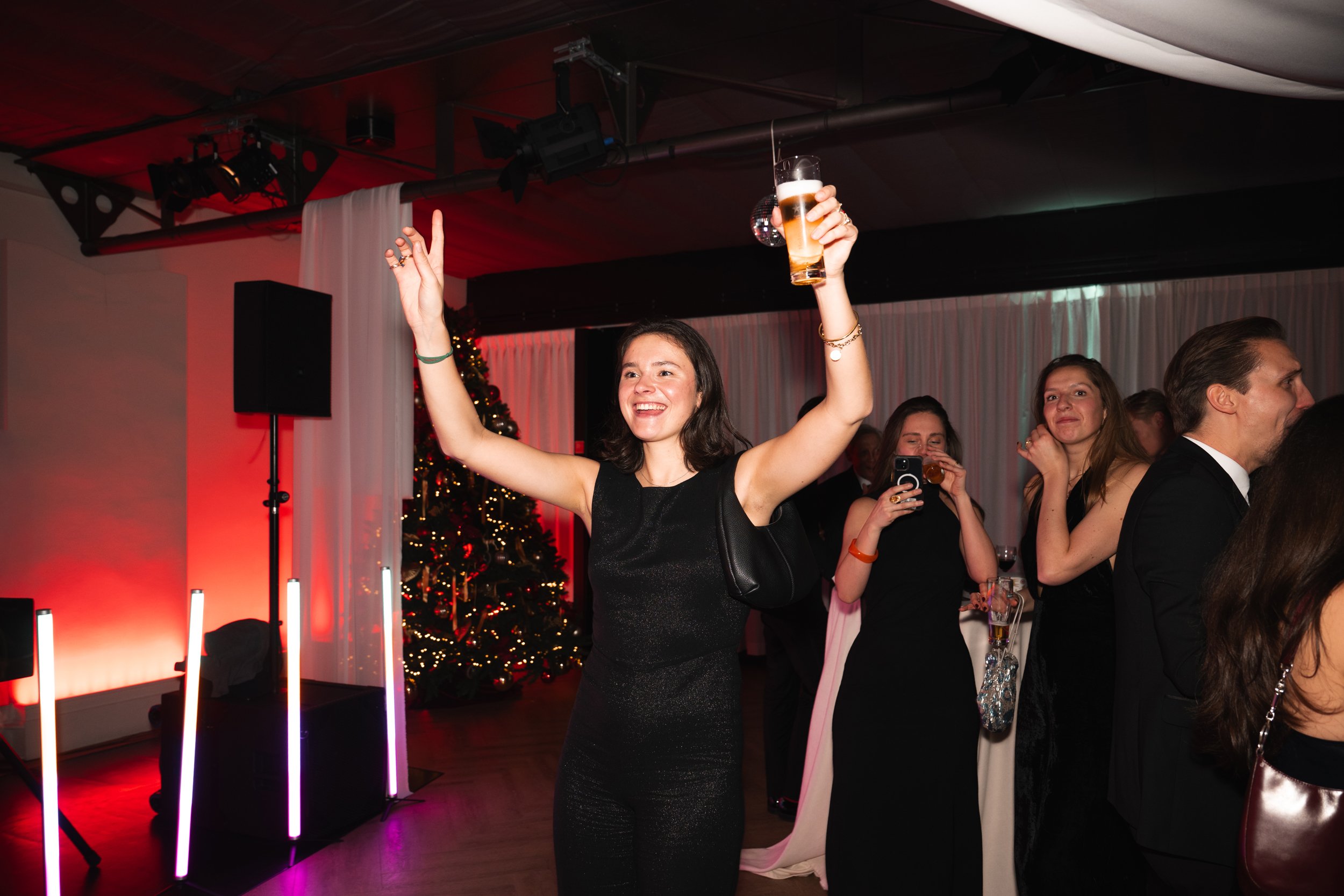 Woman celebrating at a festive party, holding a drink in one hand and raising her other hand, with Christmas decorations in the background.