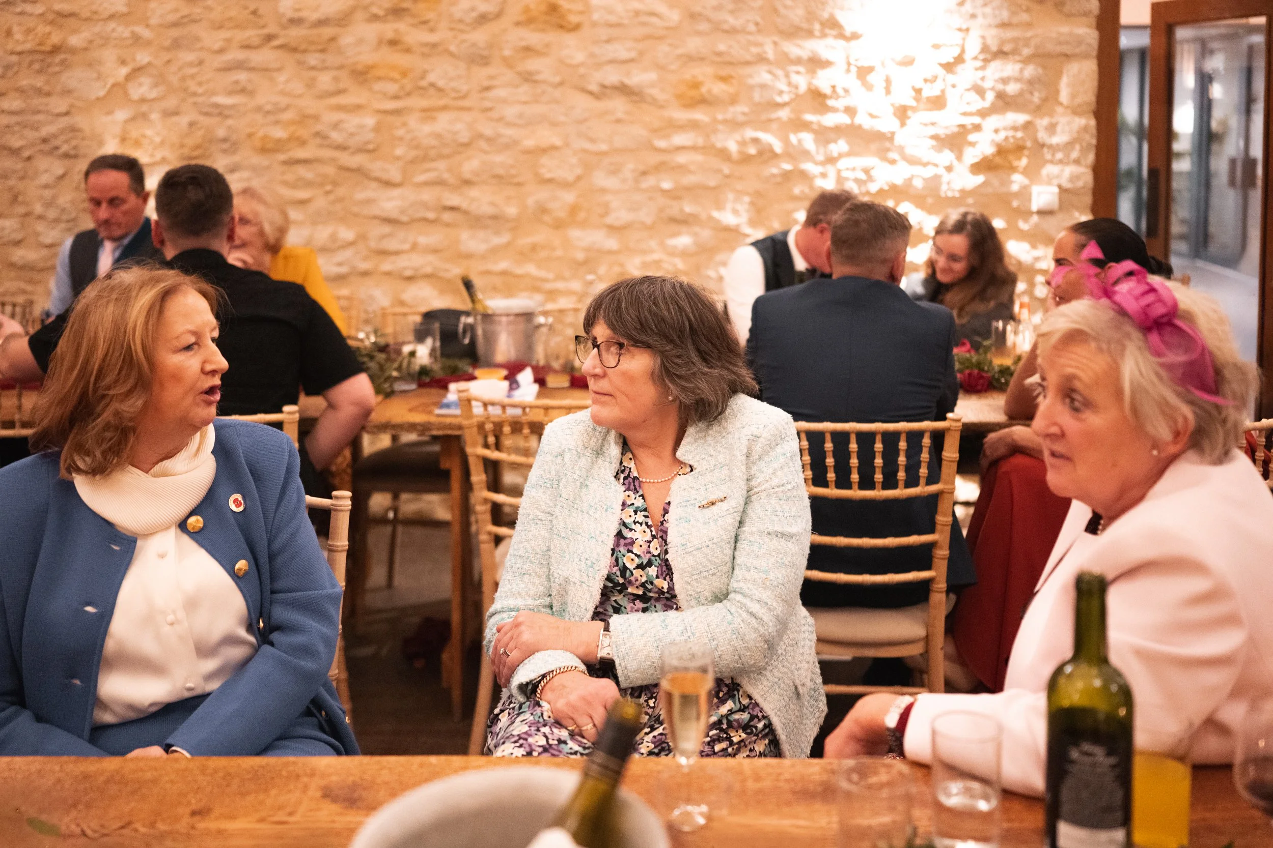 Three women engaged in conversation at a dinner event with a rustic brick wall background, surrounded by other guests and tables with wine bottles and glasses.