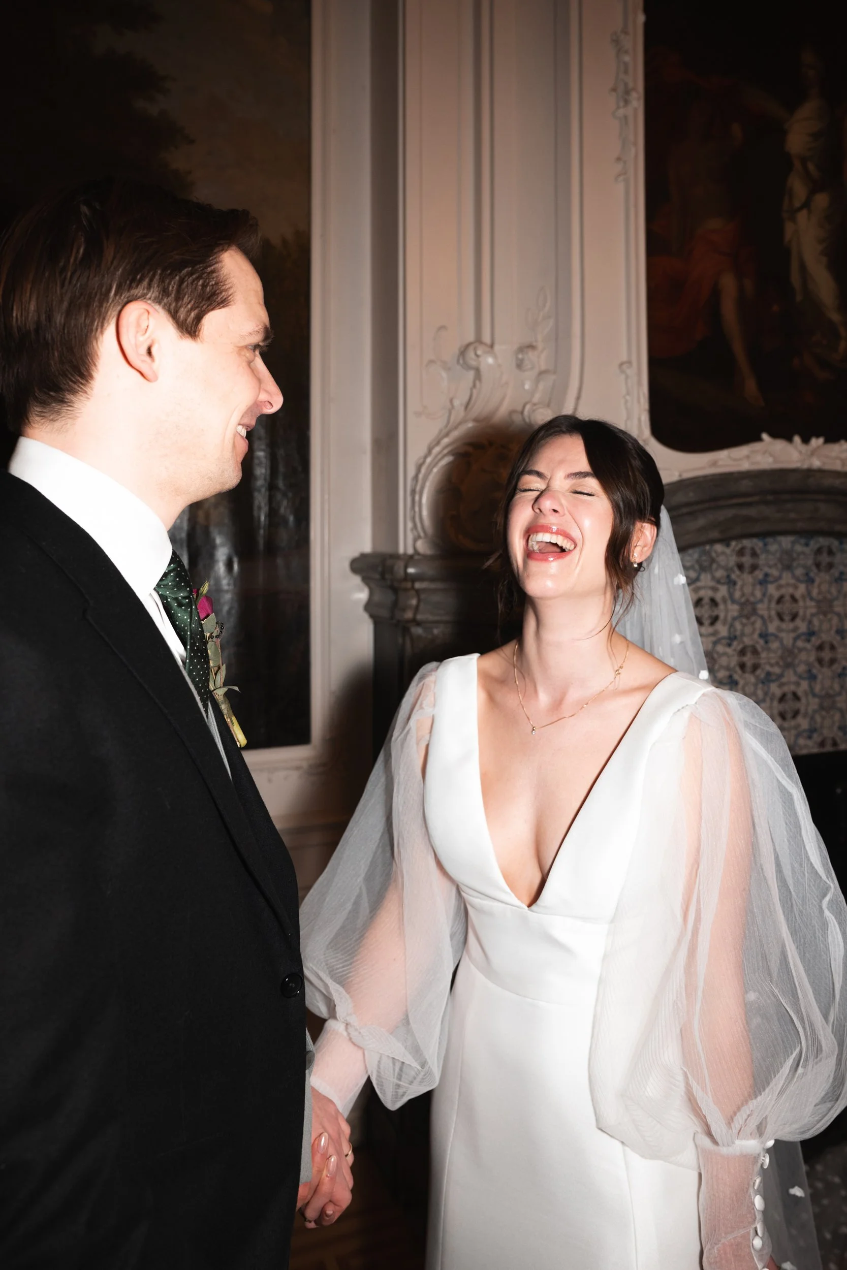 A bride and groom are holding hands and smiling during their wedding ceremony in an ornate room.