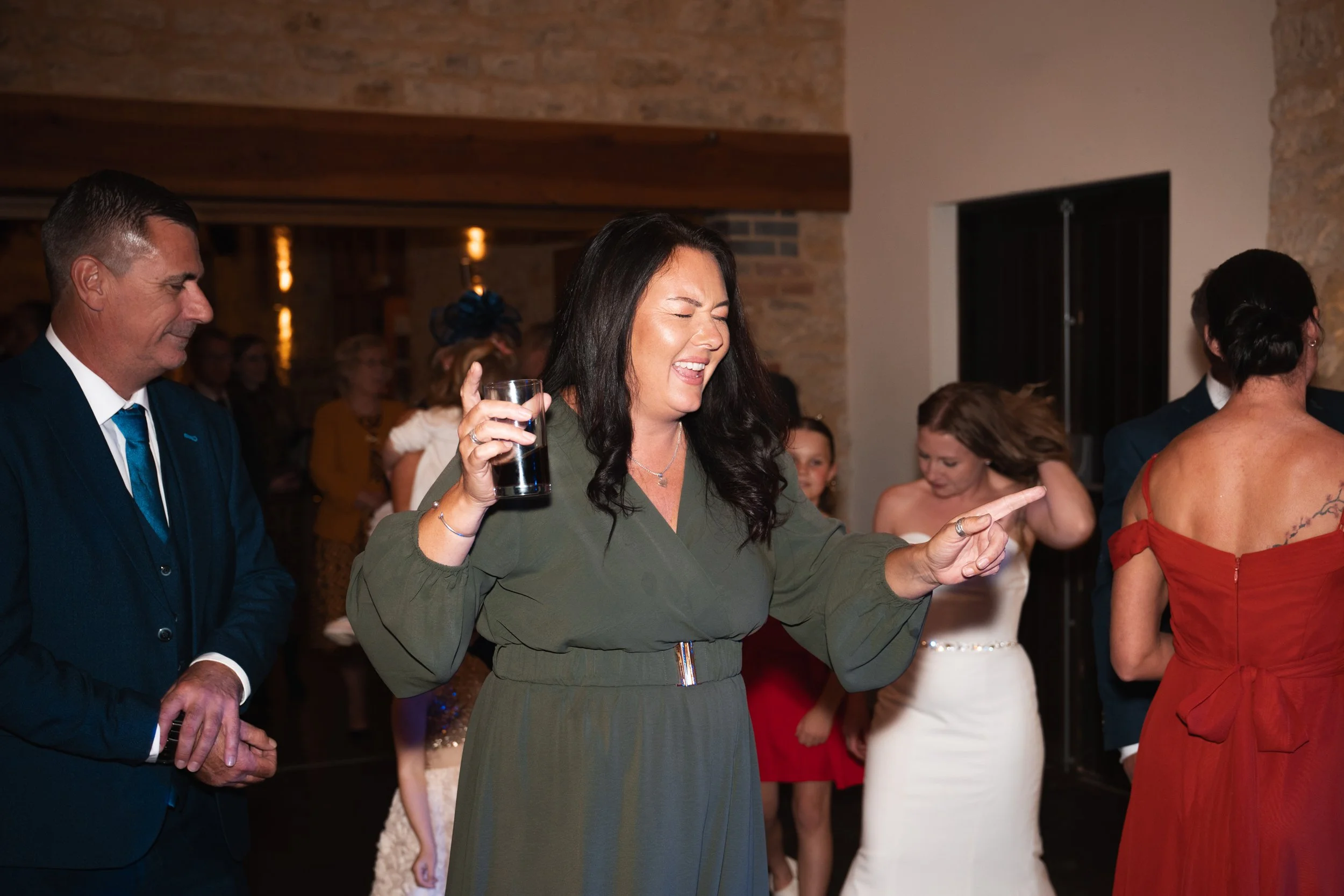 A group of people dancing at a celebration or wedding reception, with a woman in the center wearing a green dress and holding a drink, smiling and pointing.