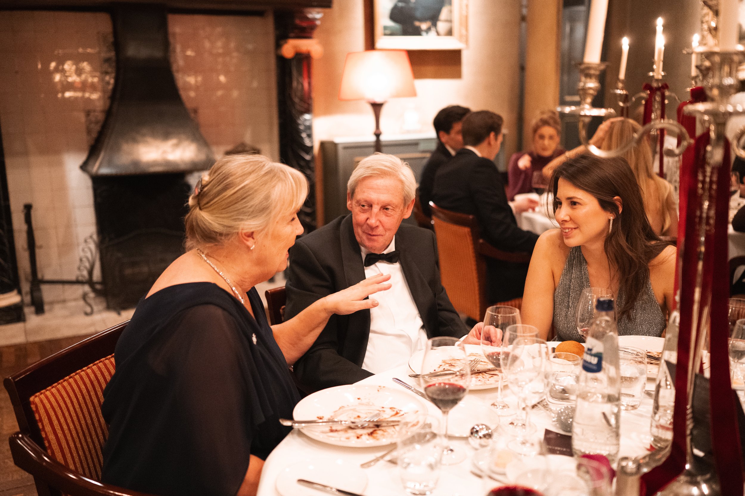 A group of people in formal attire sitting at a round dining table in a restaurant, engaging in conversation and having dinner, with wine glasses, plates, and a candelabra on the table.