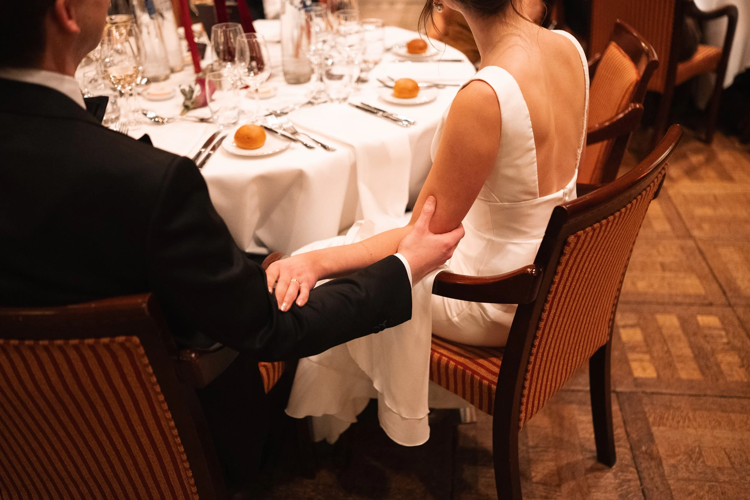 A man in a black tuxedo holding the hand of a woman in a white dress at a formal dining event.