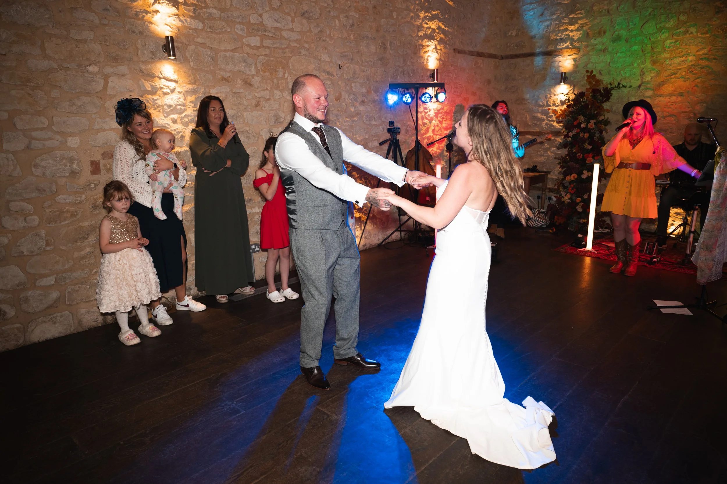A bride and groom are dancing at their wedding reception, holding hands and smiling at each other. Guests, including children, are watching nearby, and a live band is performing in the background inside a stone-walled venue.