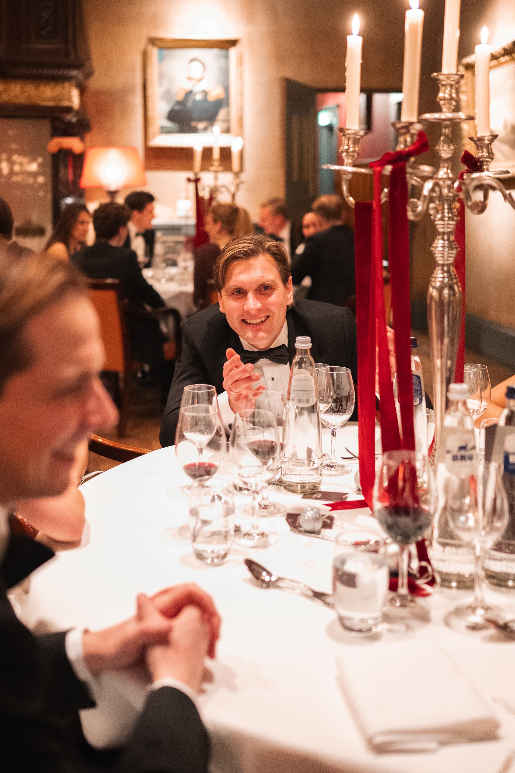 A group of people in formal attire enjoying a dinner party in a dimly lit, elegant restaurant with candles and a portrait on the wall.
