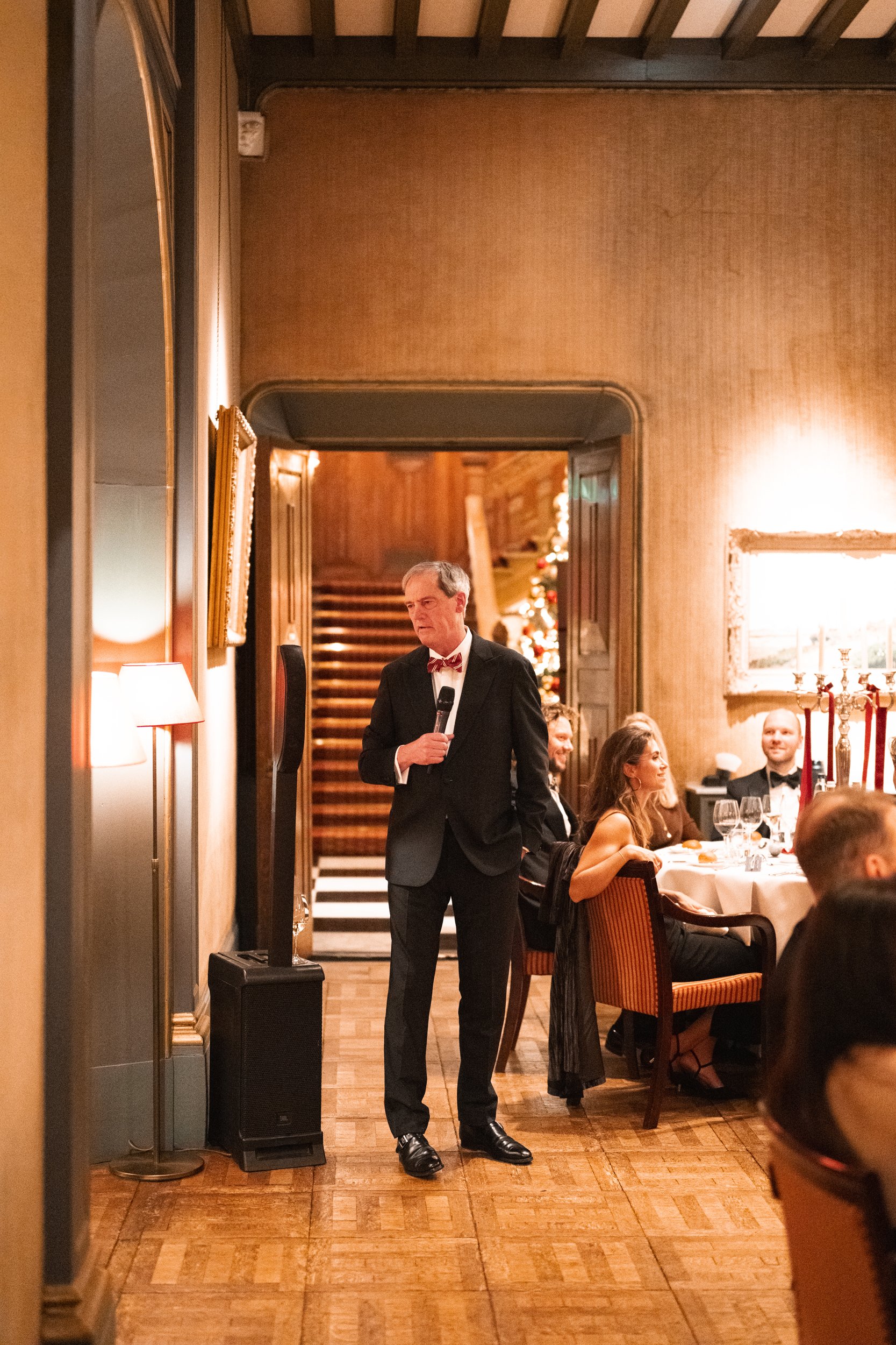 A man in a tuxedo with a bow tie holding a microphone standing at a formal dinner event with guests seated at round tables inside a warmly lit room decorated for Christmas.