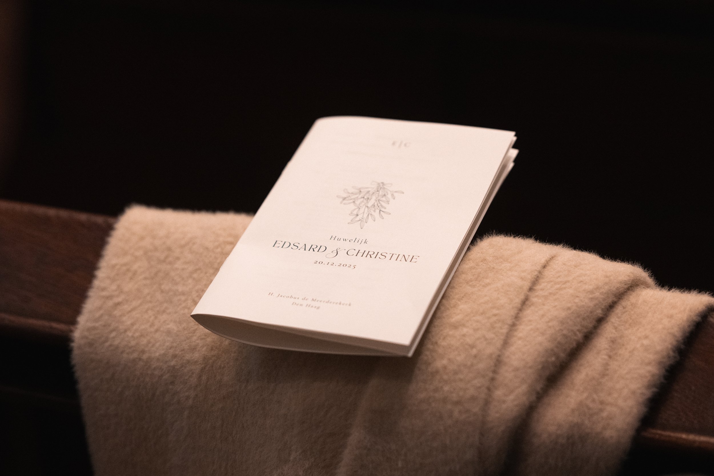 Wedding program for EDSARD & CHRISTINE on December 20, 2025, resting on a beige cloth over a wooden surface with a dark background.