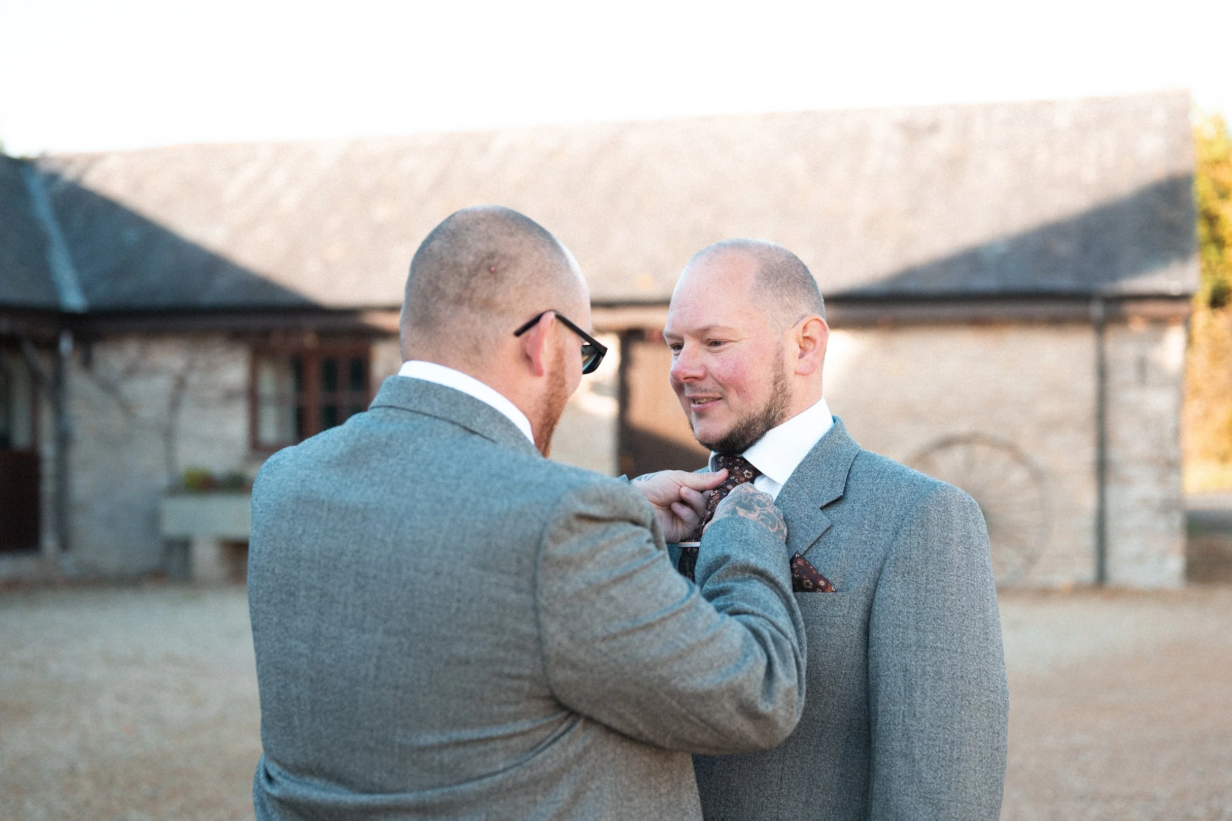 Two men in suits outdoors, one adjusting the other's tie, during daytime.