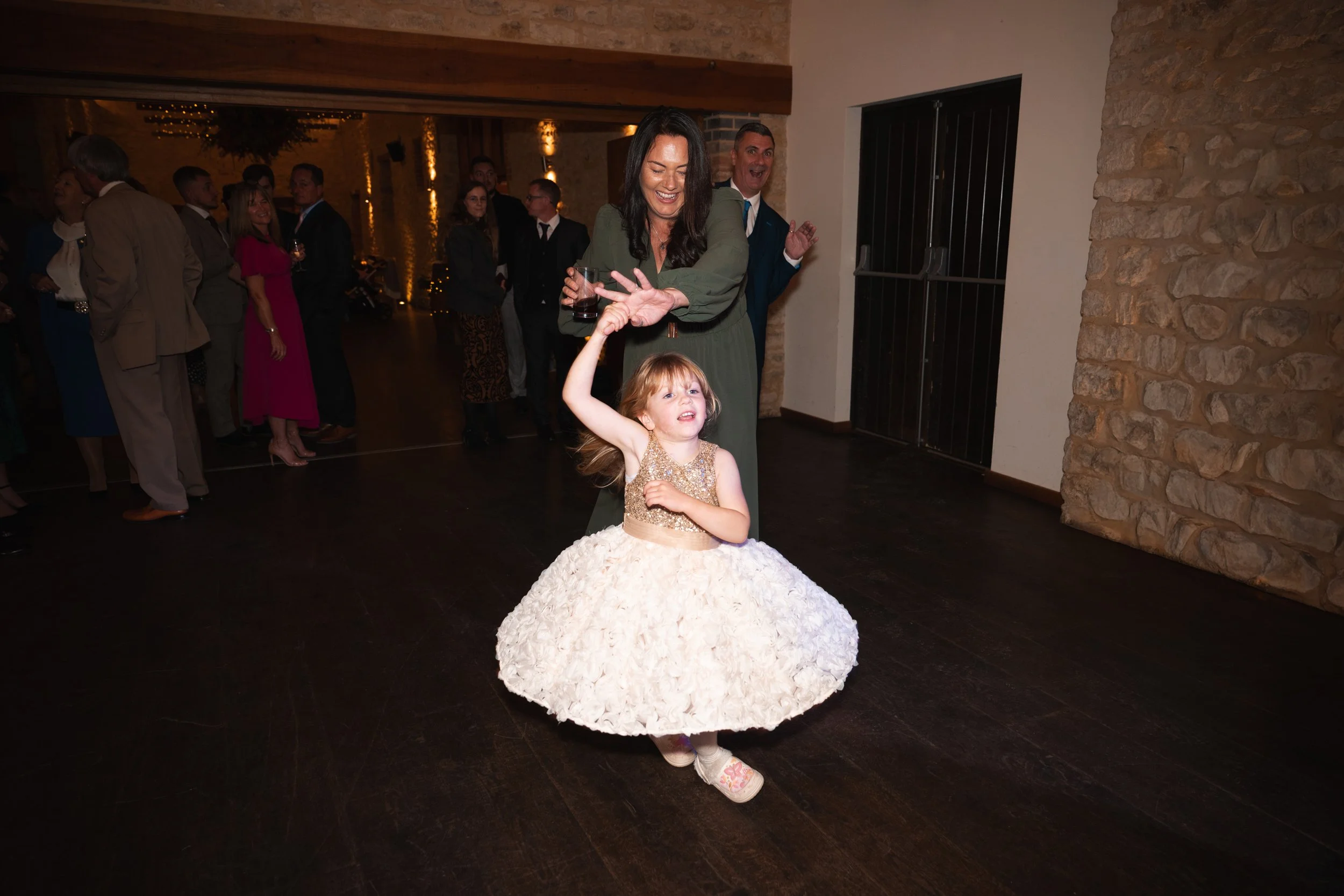 A young girl in a fancy white dress dancing with an adult woman at a party, surrounded by other guests in the background.