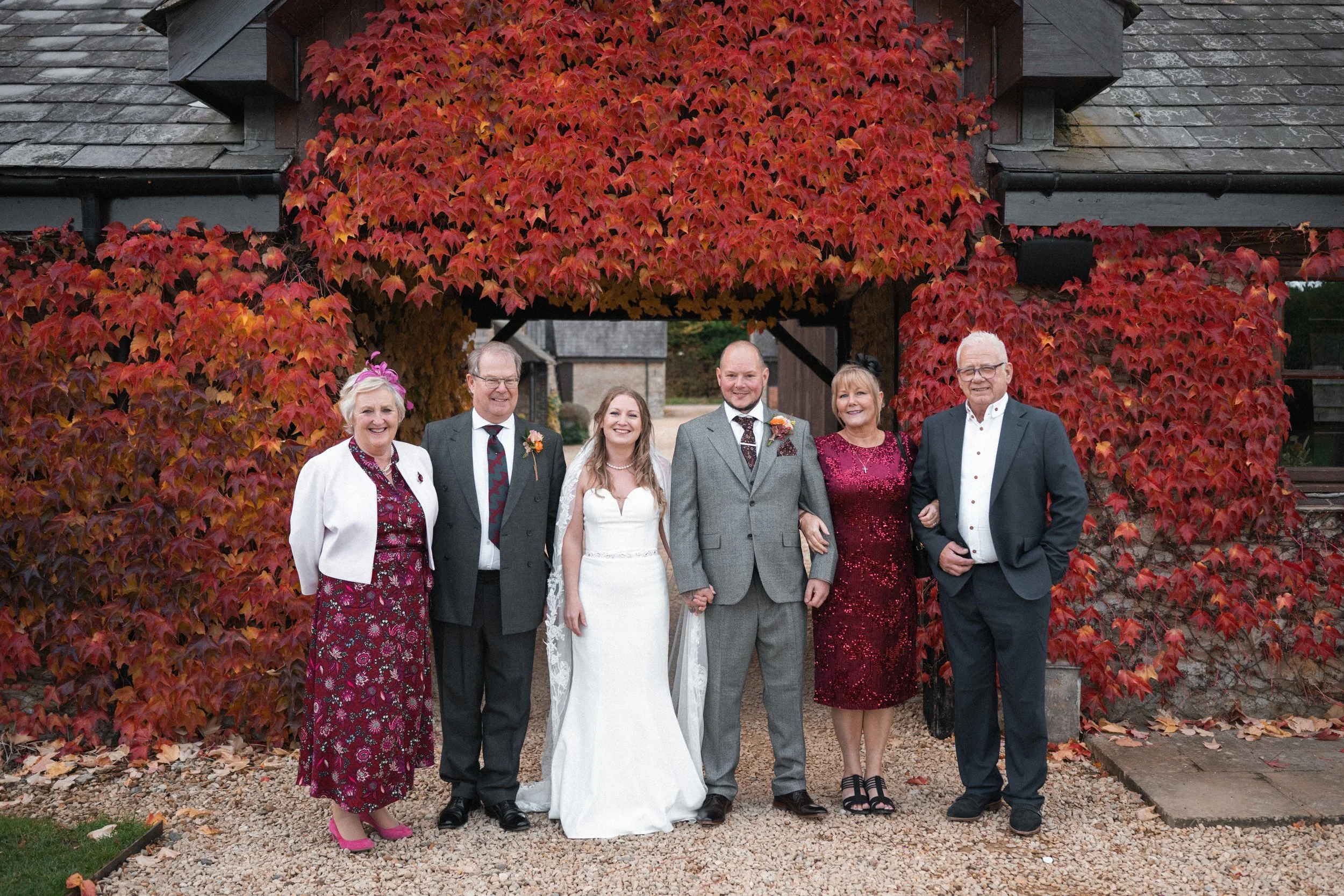 Group of seven people at a wedding, standing outdoors in front of a wooden structure with red autumn leaves. Includes bride in white gown, groom in gray suit, and five older adults in formal attire.
