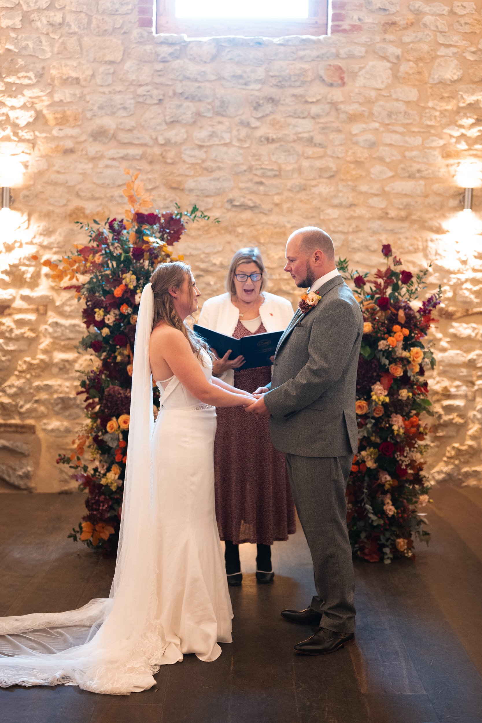 Bride and groom holding hands during wedding ceremony, with officiant standing behind them, in front of a stone wall with floral arrangements.