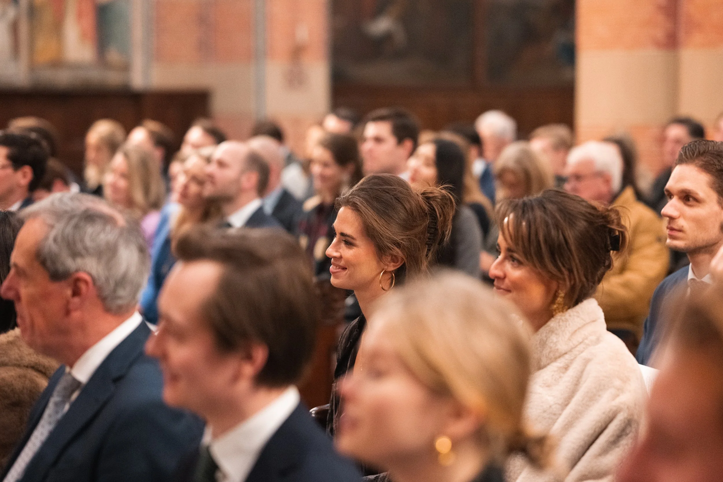 An audience seated in a hall during a formal event or conference, with people dressed in business attire, listening attentively.