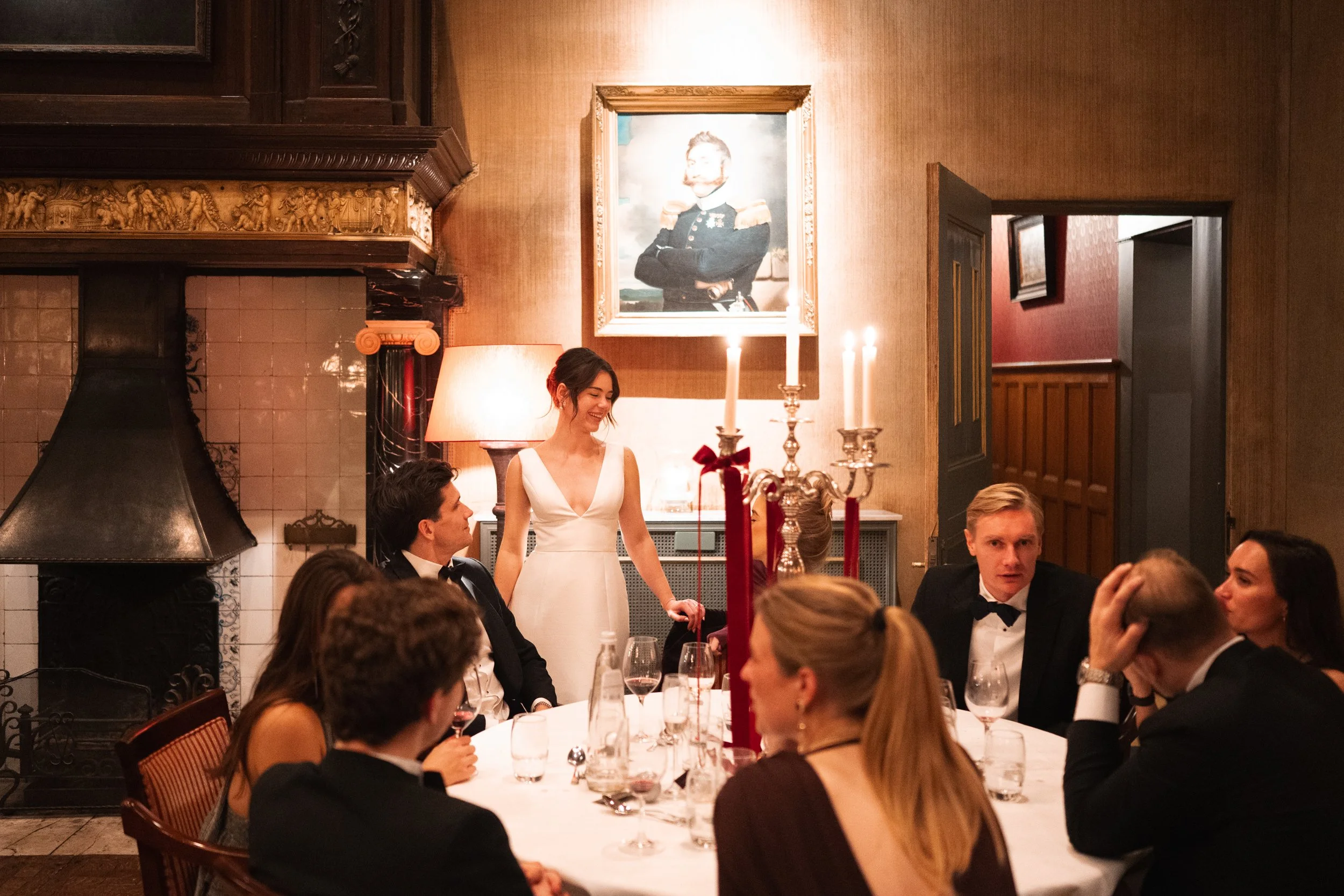 People dressed in formal attire sitting around a dining table in a warmly lit room with candles, a portrait on the wall, and a fireplace in the background.