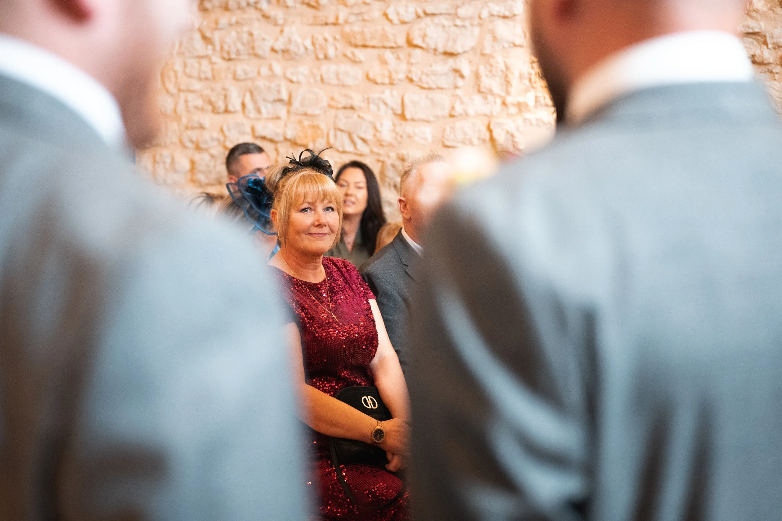 Woman in red dress with a black clutch at a formal event, sitting against a brick wall, surrounded by others in formal attire.