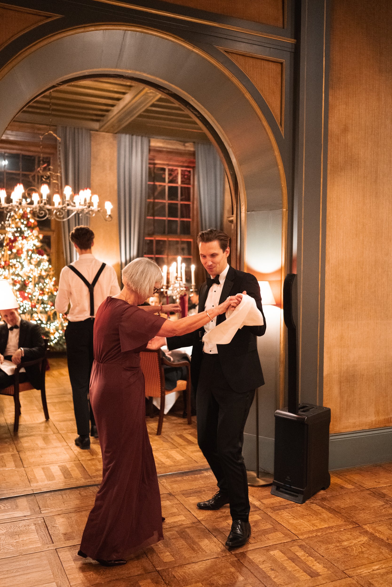 A young man in a tuxedo dancing with an older woman in a gown at a holiday celebration in an elegant room decorated with a Christmas tree, chandeliers, and candelabras.