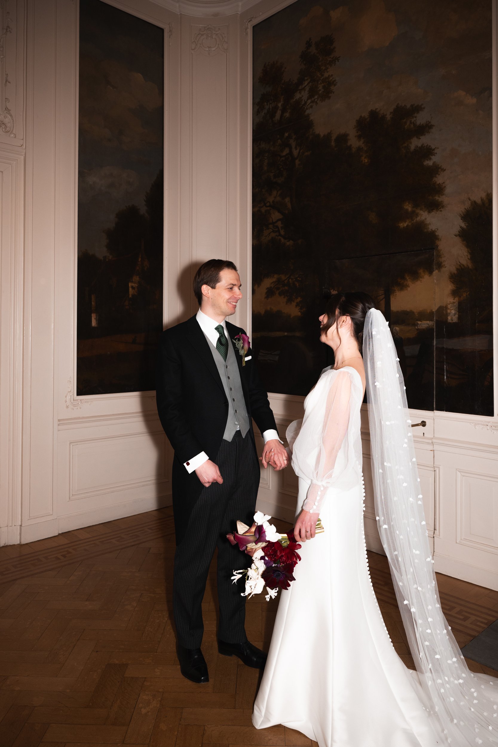 A bride and groom holding hands and smiling at each other indoors, with the bride holding a bouquet of flowers, during their wedding ceremony.