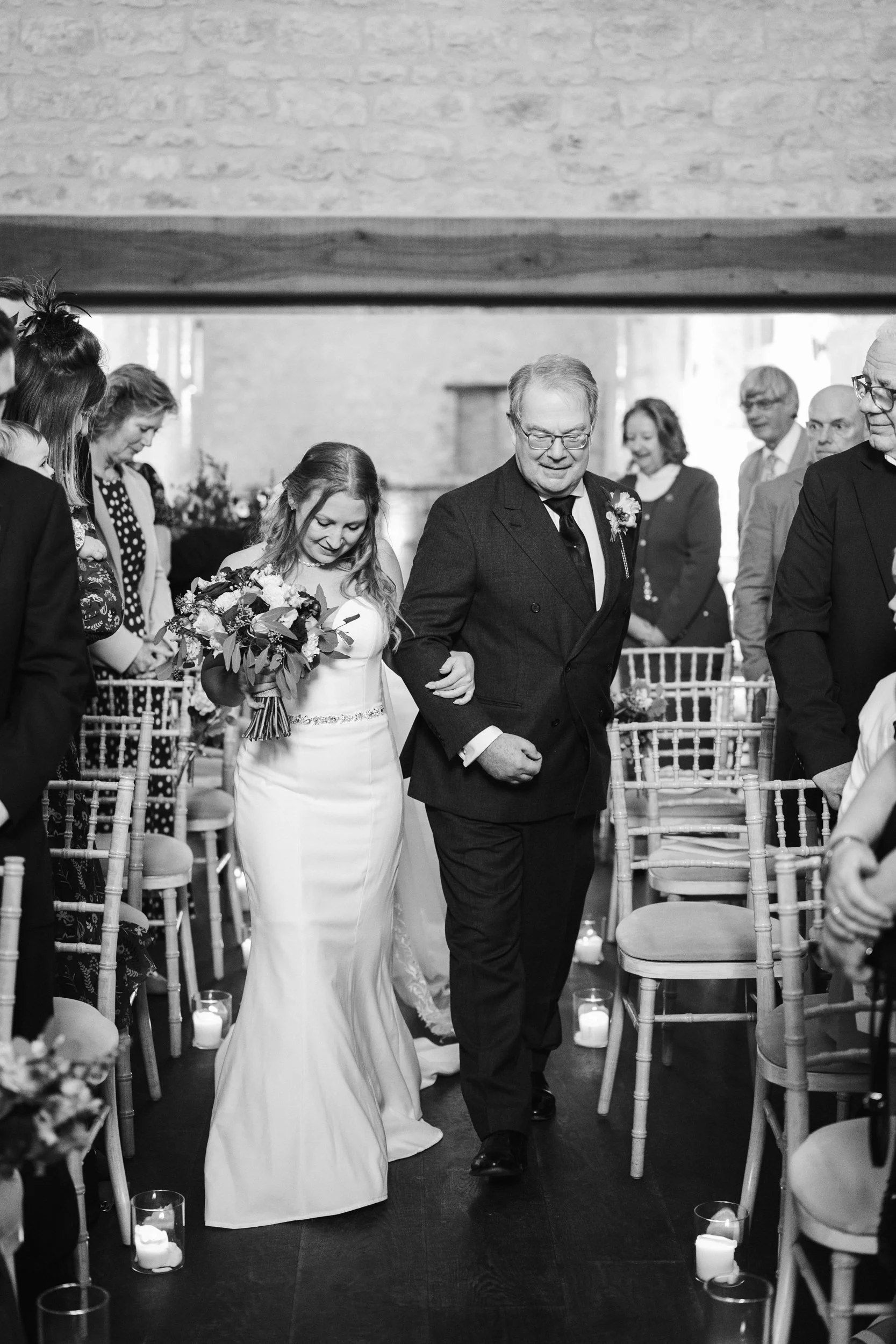 A bride walking down the aisle with her father during a wedding ceremony in a church, surrounded by guests seated on either side.