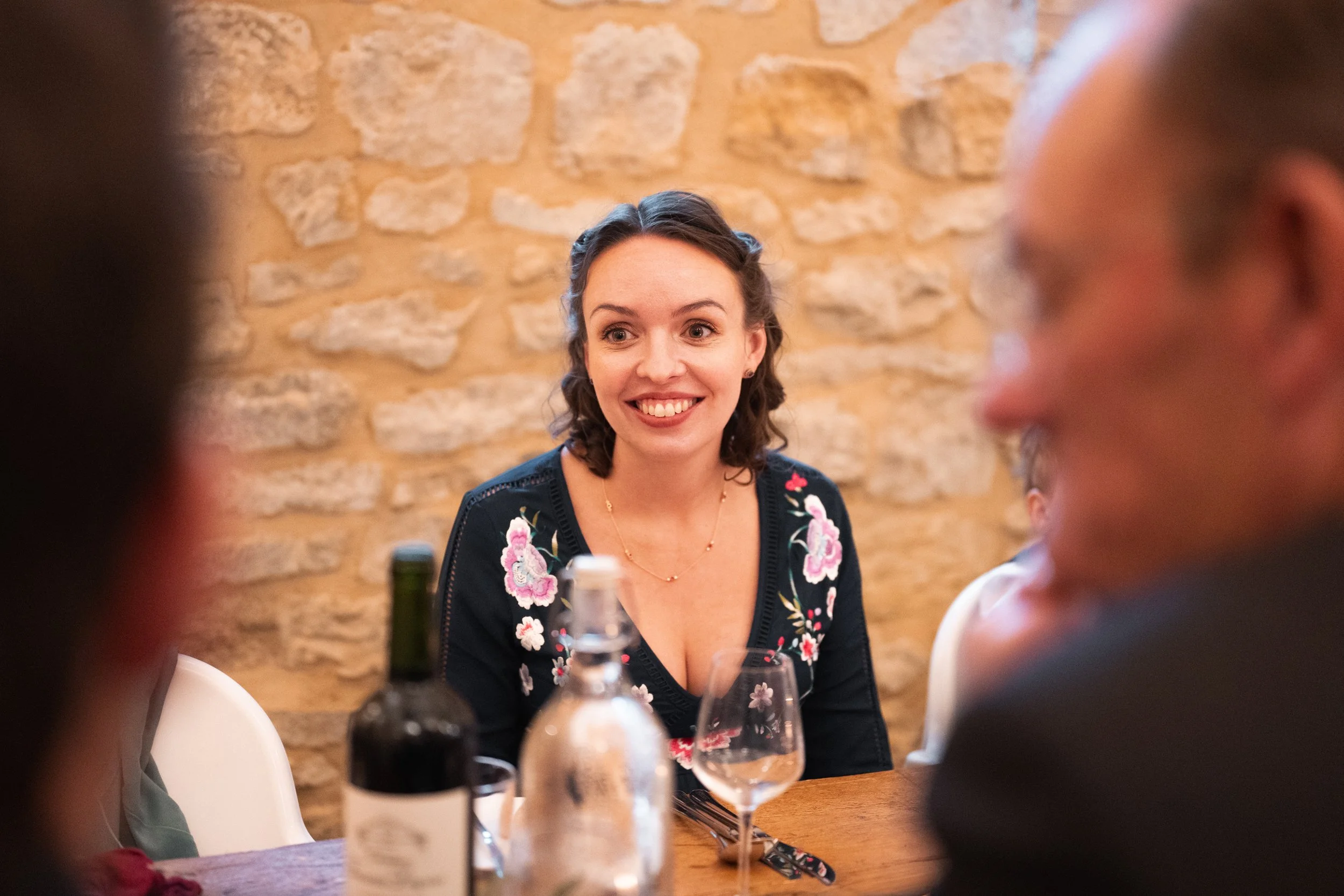A woman smiling at a dinner table with a wine glass, a bottle of wine, and a water bottle, surrounded by blurred people, with a stone wall background.