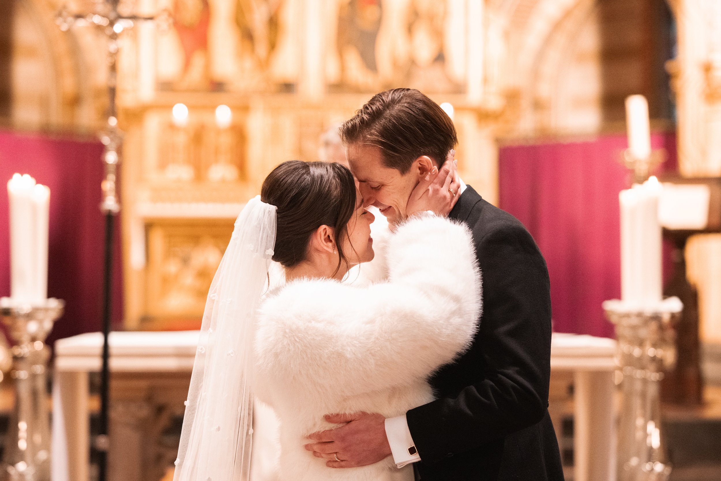 A newlywed couple embracing in a church, sharing a romantic moment with foreheads and noses touching. The bride is in a white wedding dress with a veil and a white fur stole, while the groom is in a black suit. They are smiling softly with eyes close