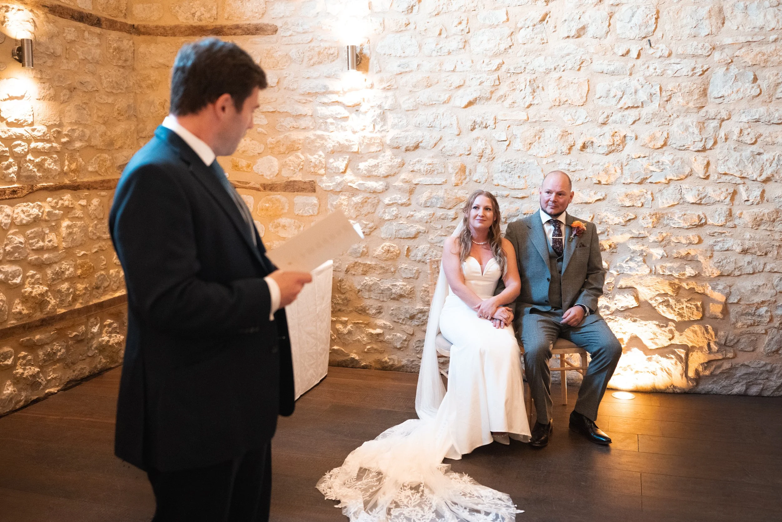 A wedding ceremony with the bride and groom sitting against a stone wall, while a man reads from a paper in front of them.