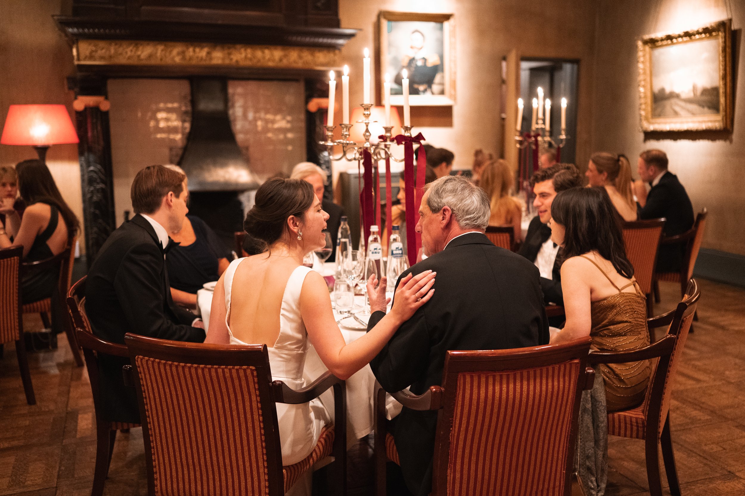 People in formal attire dining in a warmly lit restaurant with a fireplace, paintings, and a candelabra on the table.