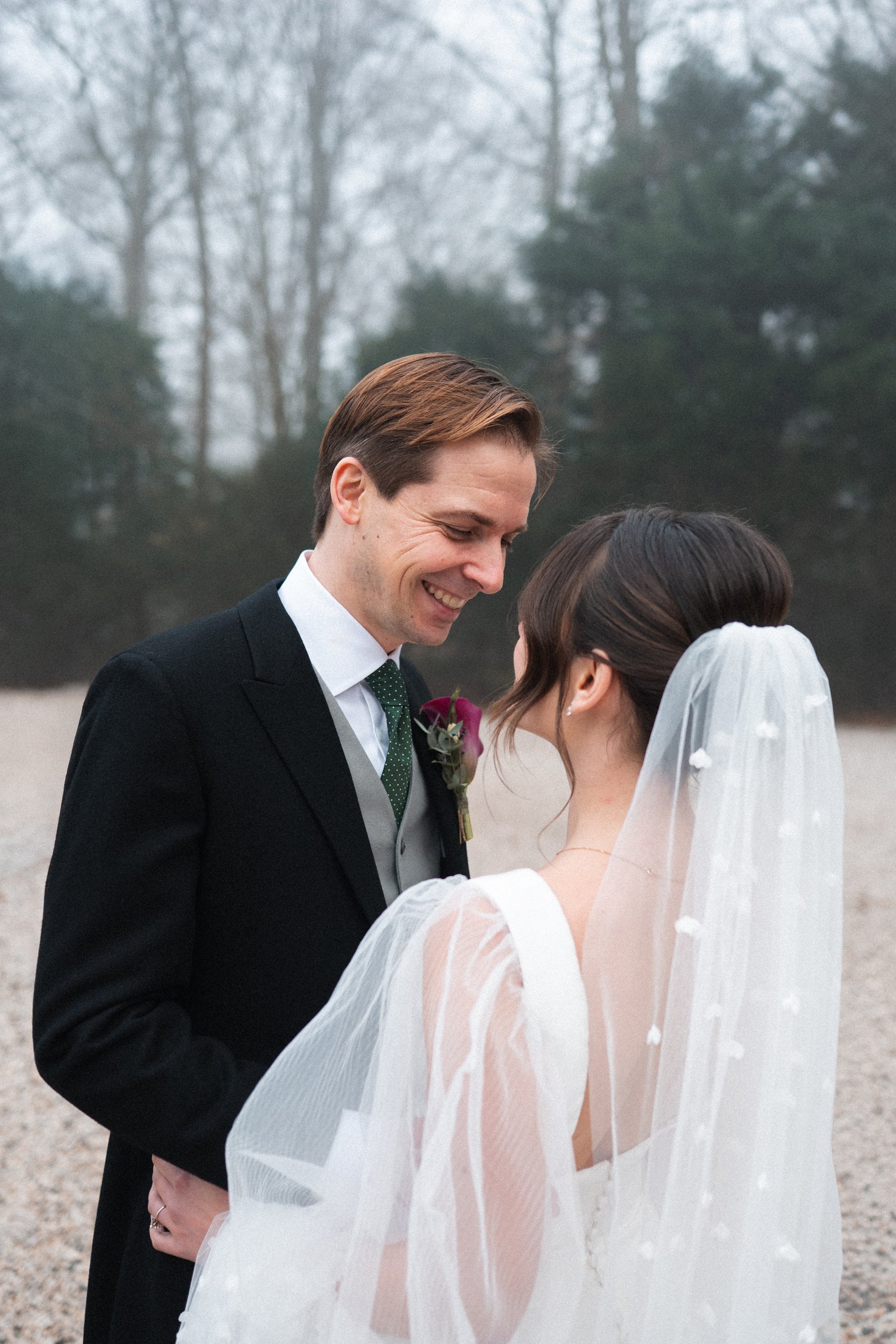 A bride and groom smiling and gazing at each other outdoors on a cloudy day, with trees in the background.