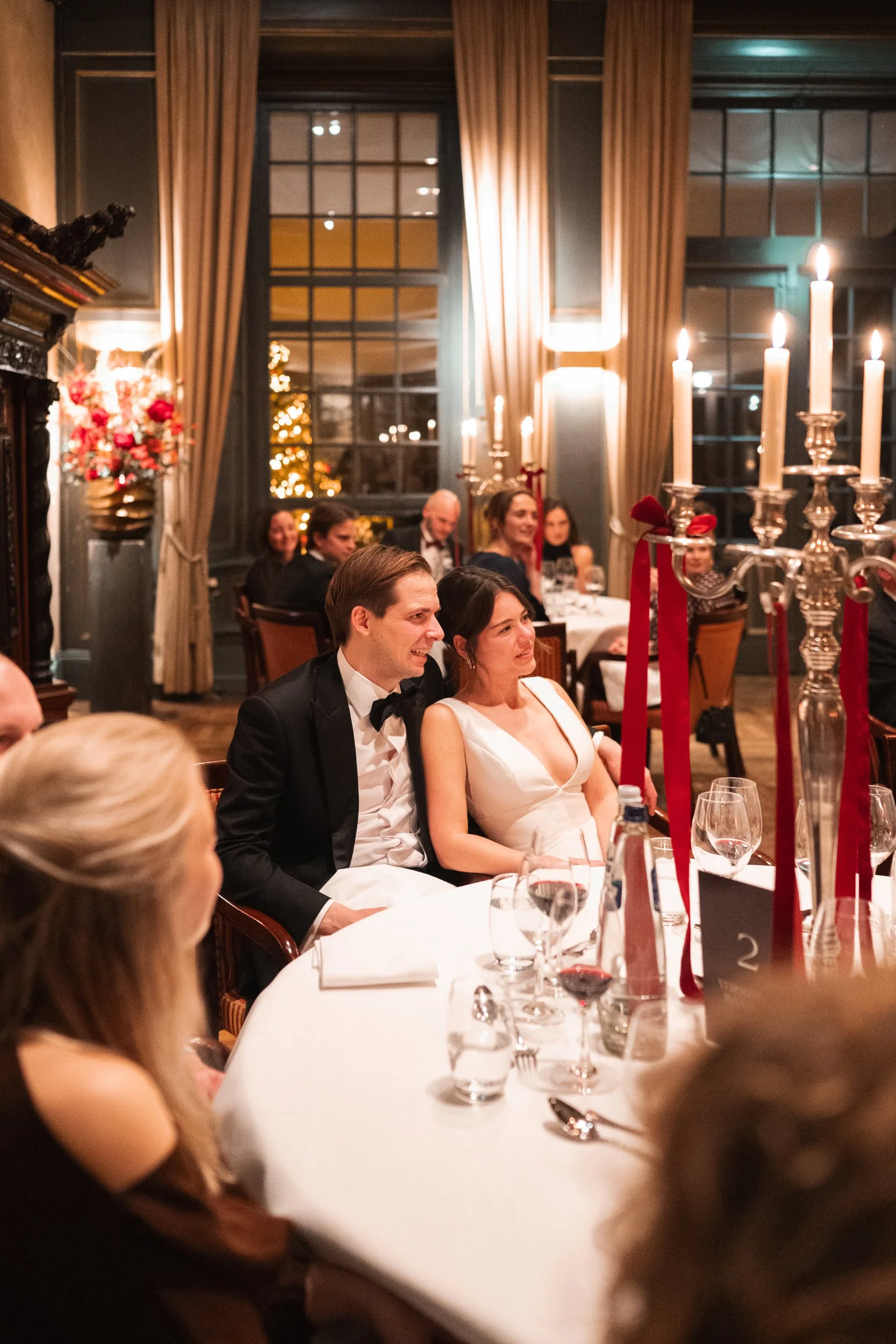 A wedding reception with a bride and groom sitting at a table, smiling and listening to a speech, surrounded by elegant decorations, candles, and guests in a warmly lit, decorated room.