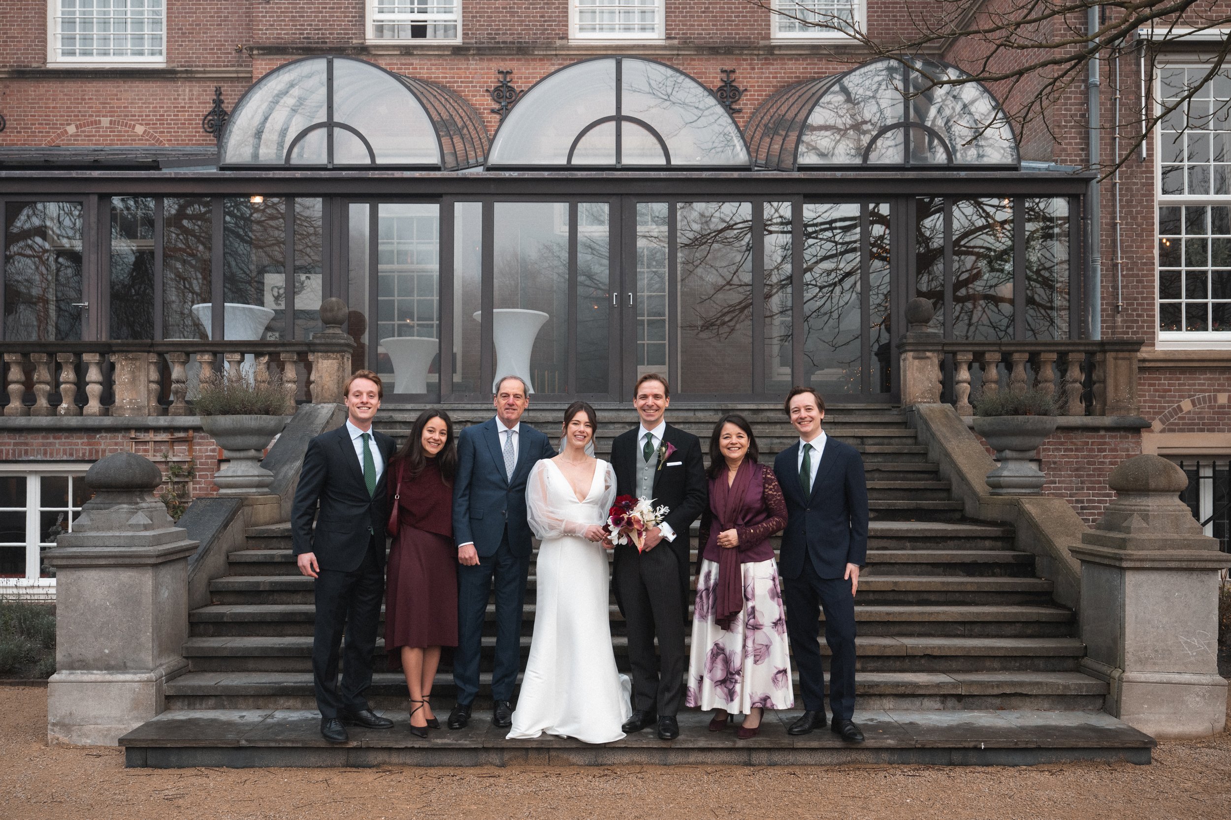 Group of seven people dressed in formal attire standing on a stone staircase in front of a brick building with glass conservatory.