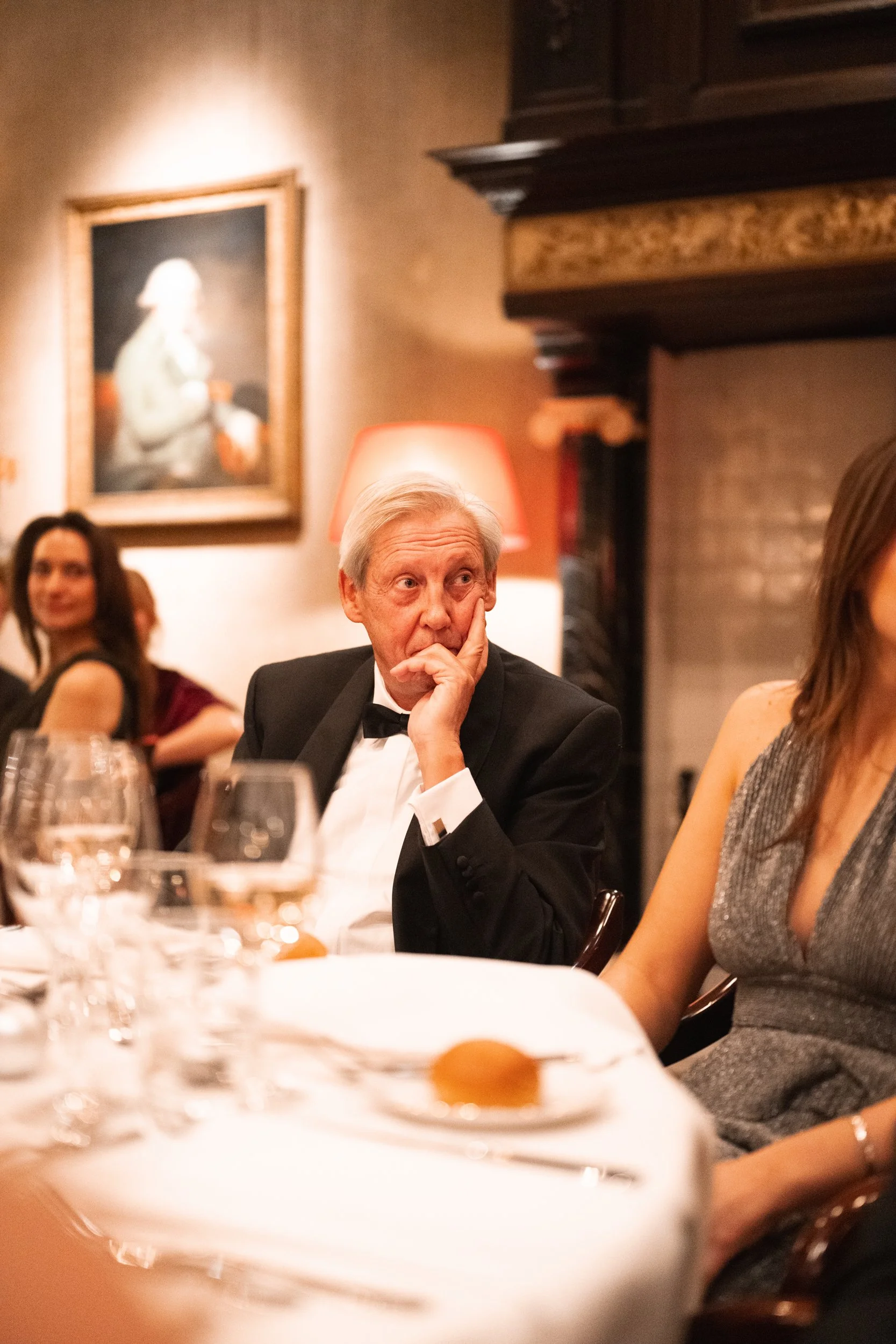 An elderly man in a tuxedo sitting at a formal dinner table, resting his chin on his hand, with a woman in a silver dress beside him at an elegant gathering.