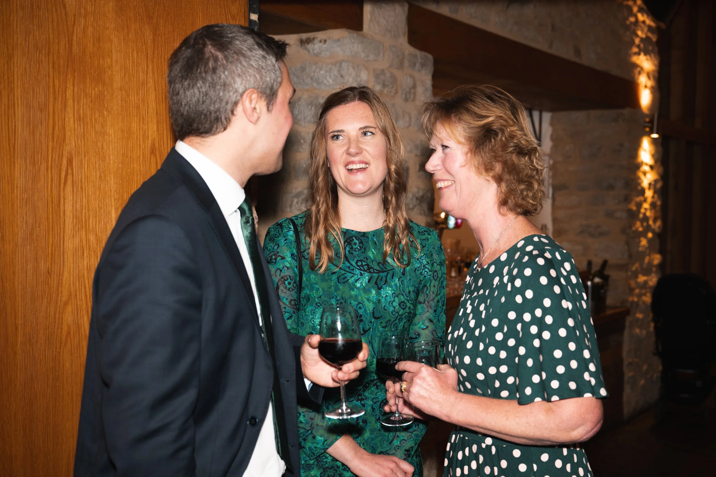 Three people at a social gathering, two women and one man, holding glasses of red wine, engaged in conversation in a warmly lit room with stone walls and wooden accents.